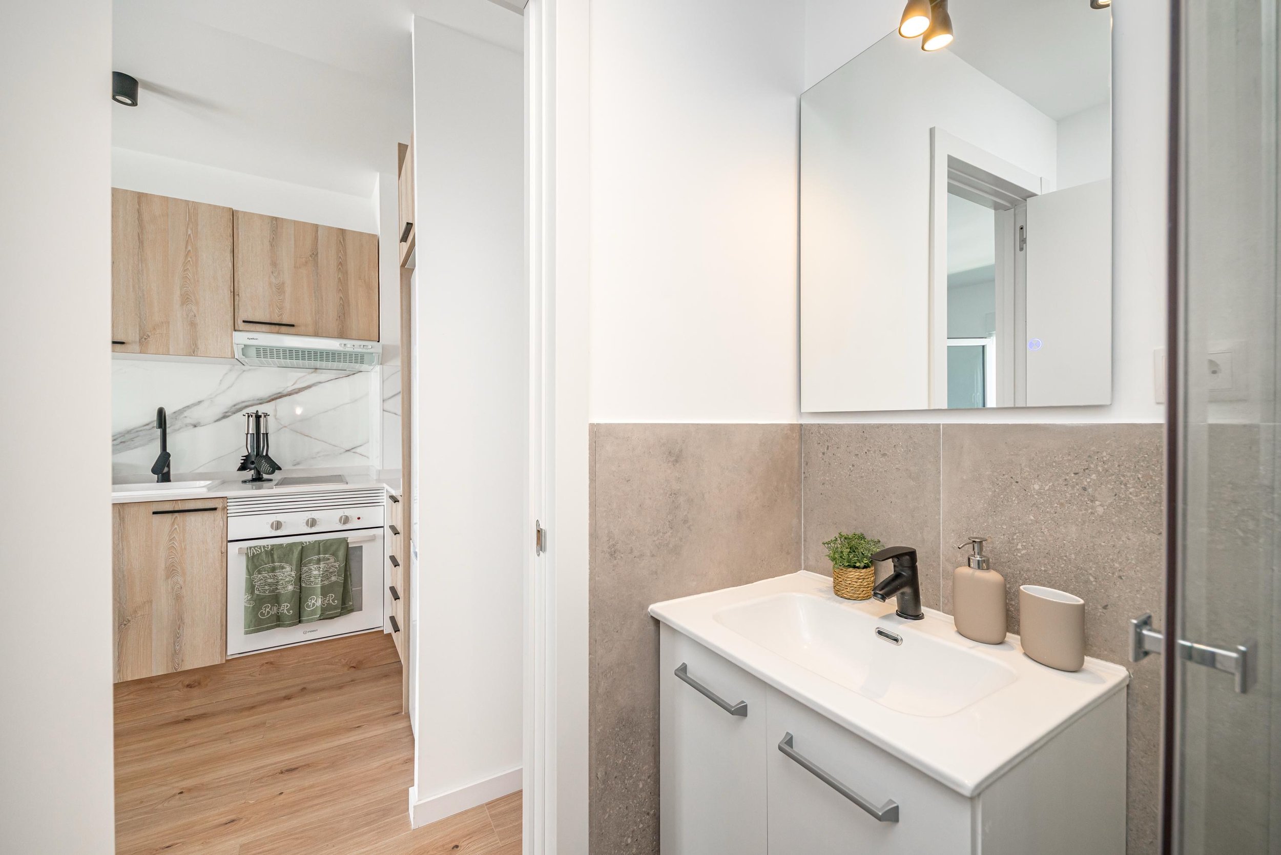 Baño con lavabo, espejo grande, artículos de tocador y una vista parcial del área de la cocina con gabinetes de madera y estufa.