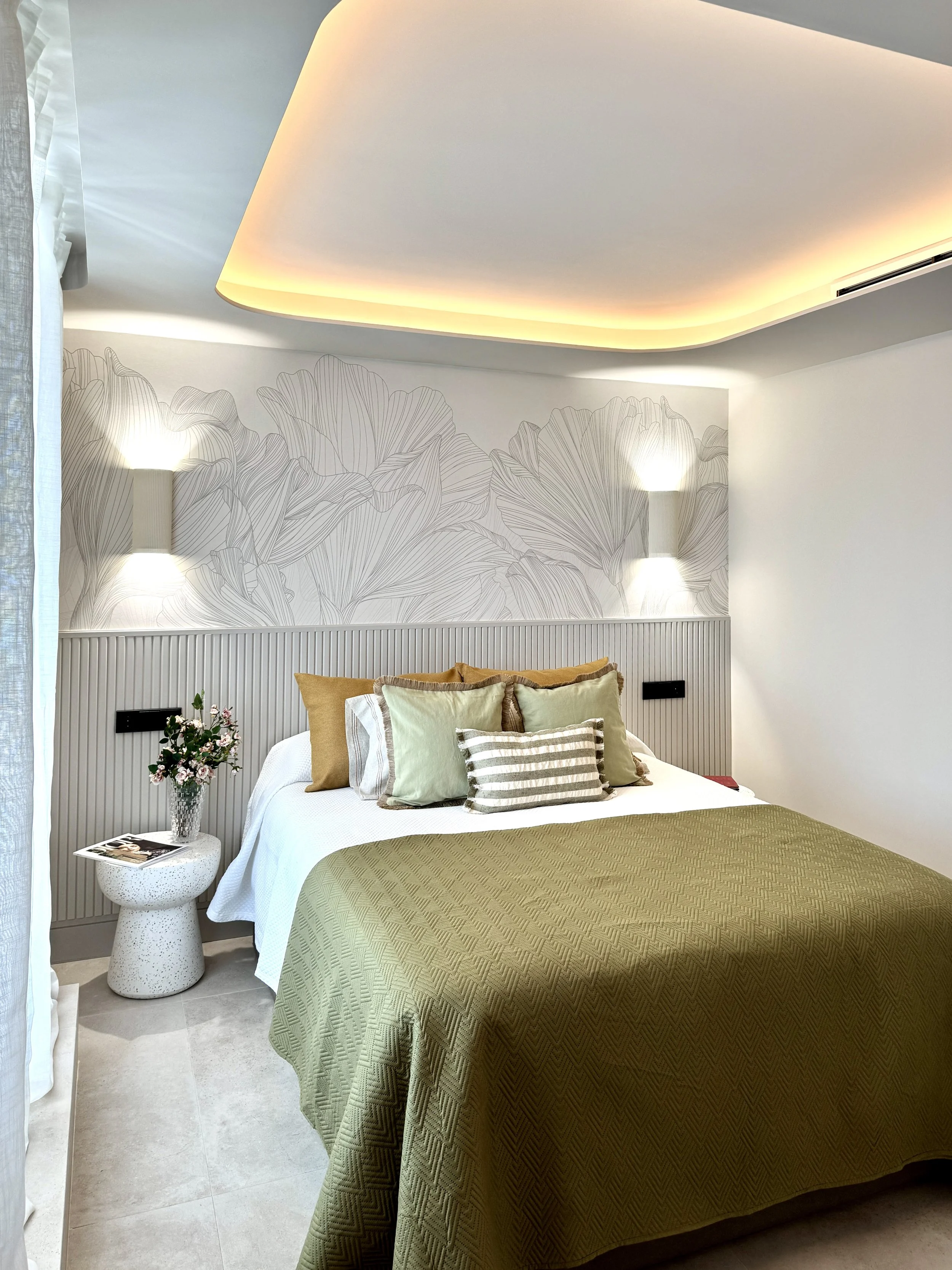 Habitación moderna con cama decorada con varias almohadas en tonos beige, blanco y gris, colcha verde, cabecera de listones blancos, paredes blancas con un mural de líneas de hojas y luces de pared. Al lado hay una mesa con una flor y revistas.