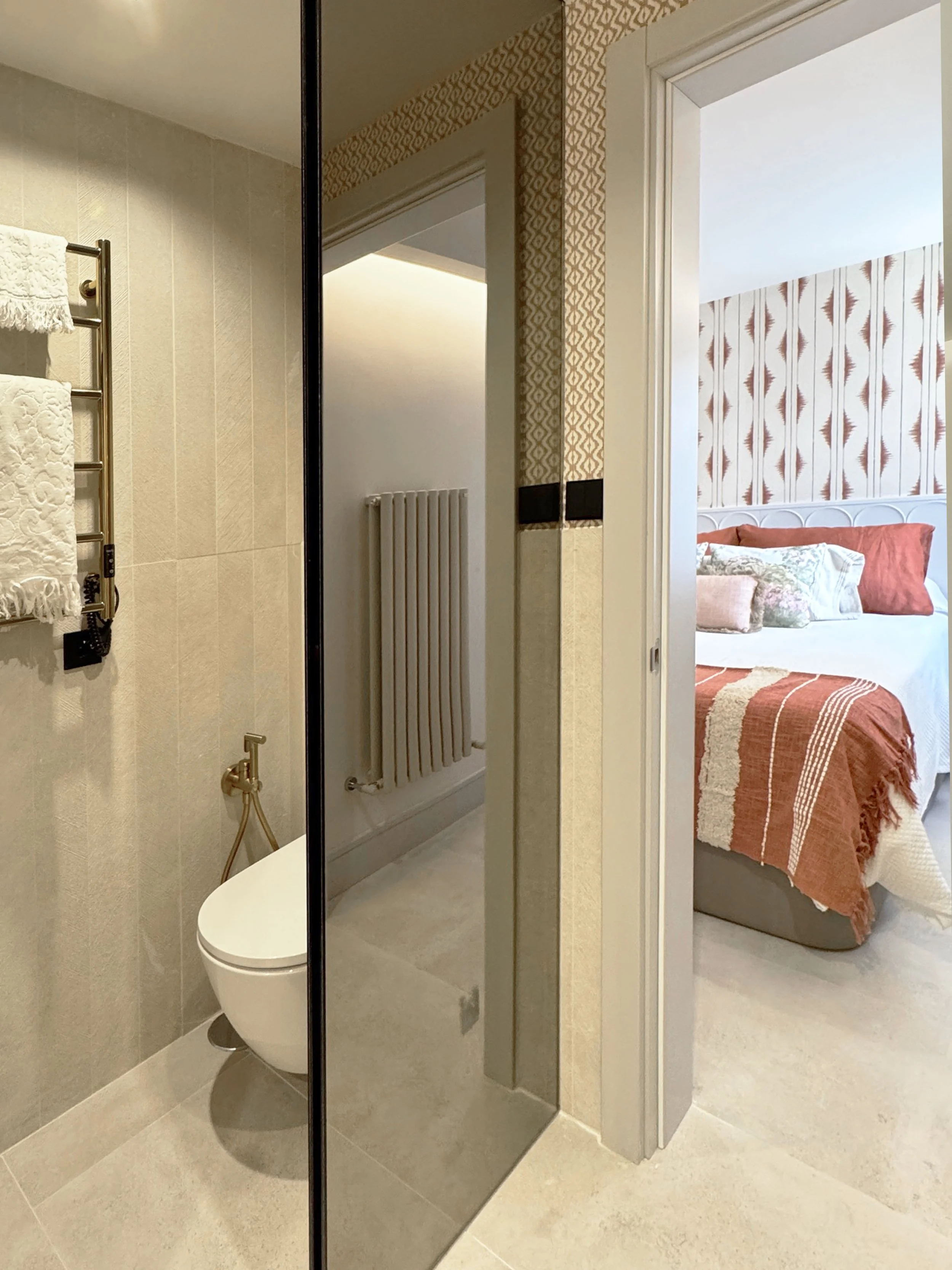 Cuarto de baño visible con espejo grande y ventana que muestra una habitación con cama decorada con cojines y cobertor en tonos terracota y beige, paredes con papel tapiz con diseño de hojas en tonos marrón y beige.