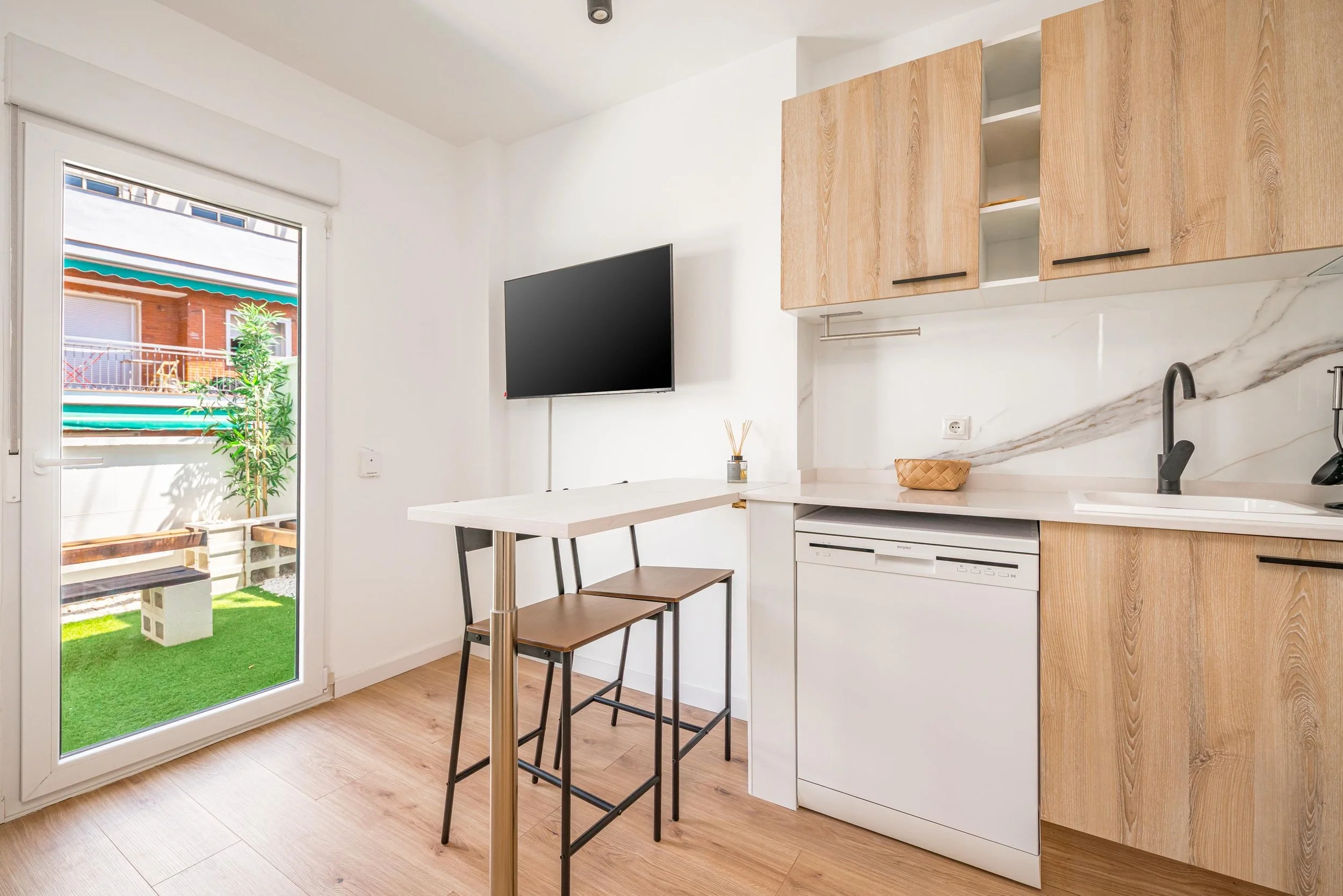 Cocina moderna con puertas de madera y electrodomésticos blancos, vista a un balcón con plantas y muebles pequeños.