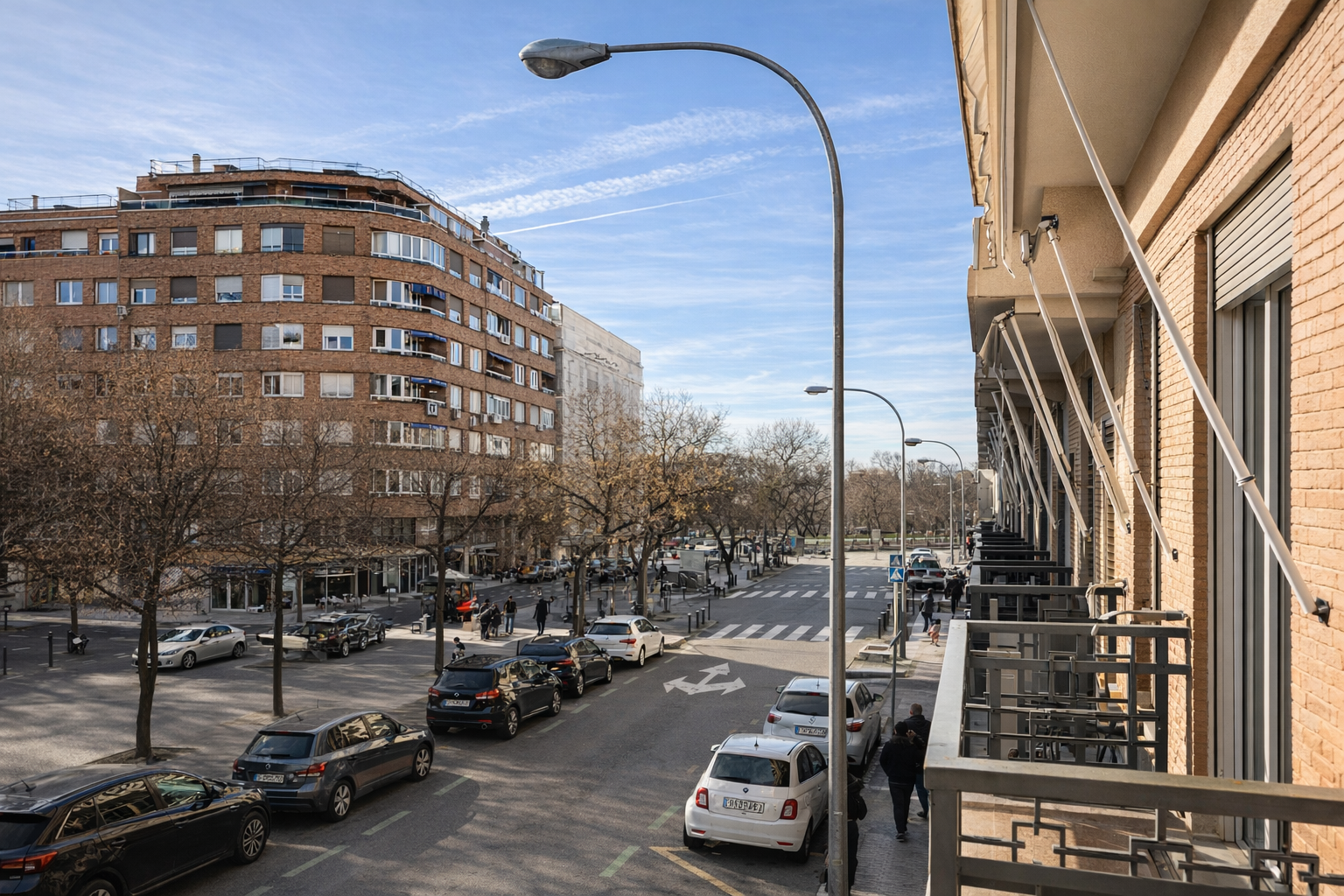 Vista de una calle en una zona urbana con edificios de apartamentos, árboles sin hojas, autos estacionados y personas caminando, en un día soleado con cielo parcialmente nublado.