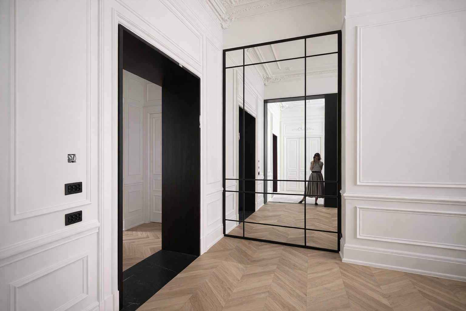 Interior de un apartamento con paredes blancas y suelo de madera en espiga, con un espejo grande de estructura negra que refleja una mujer que lleva una falda larga y camiseta, y varias puertas blancas con molduras.