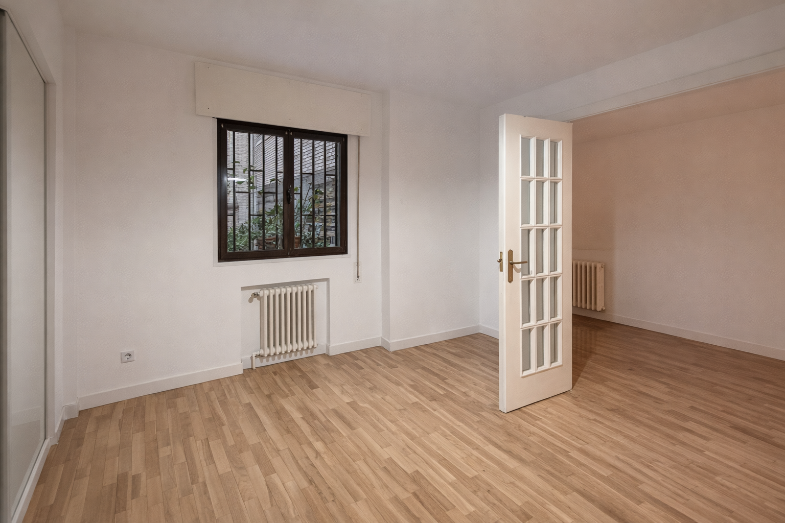 Habitación vacía con paredes blancas, piso de madera, ventana con reja y radiador, puerta con vidrio