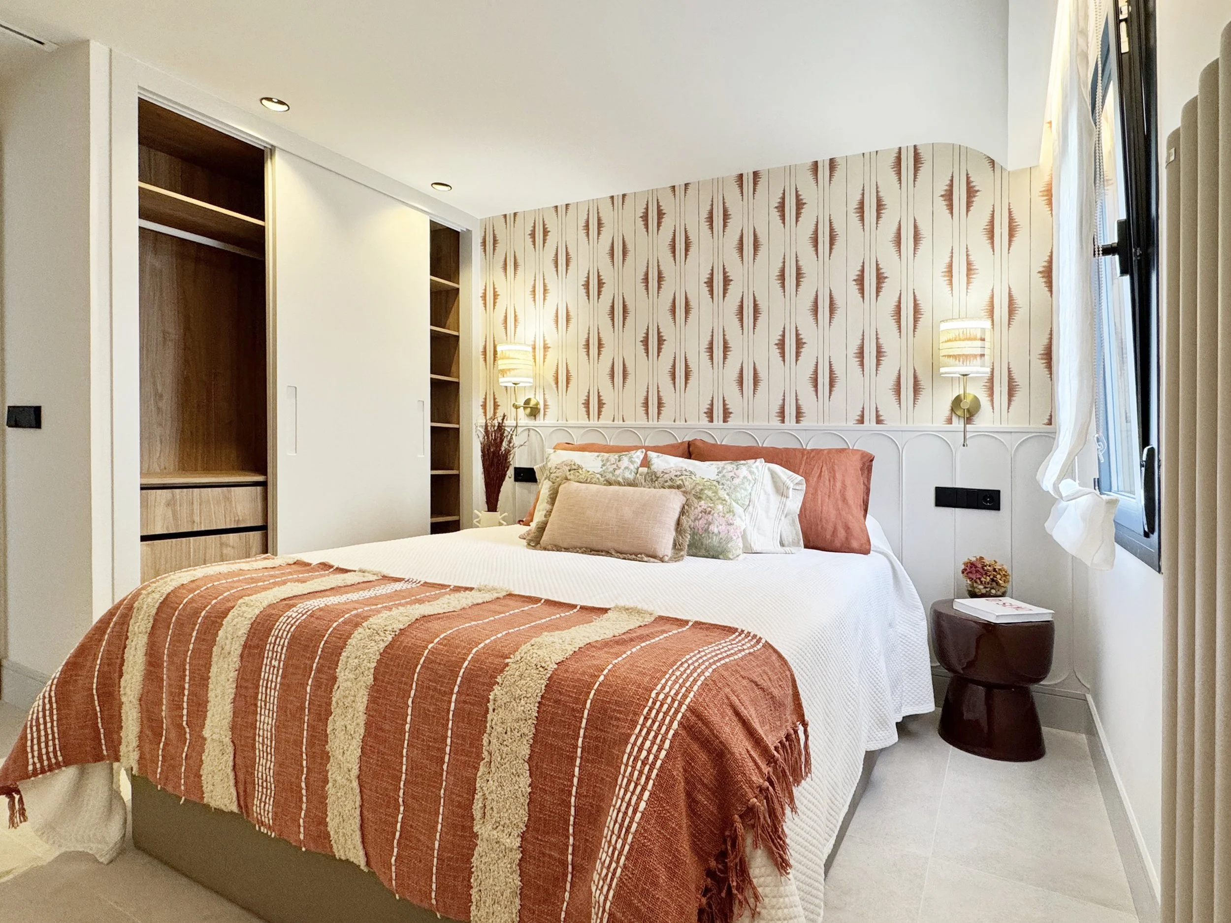 Habitación con cama grande, ropa de cama blanca, múltiples cojines de diferentes colores y diseños, pared decorativa con papel tapiz de patrón geométrico en tonos naranjas y beige, lámparas de pared, ventana con cortina blanca, mesita de noche con li