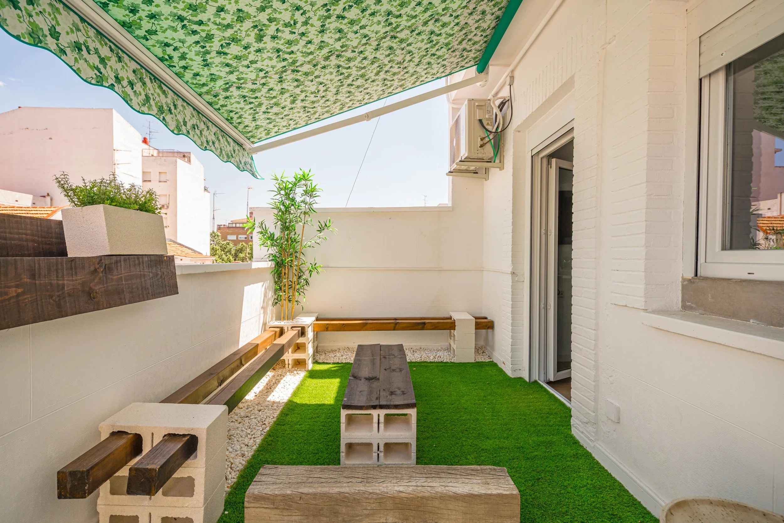 Balcón con césped artificial, plantas, bancos de madera y muro de ladrillos blancos, con toldo verde con pattern de hojas.