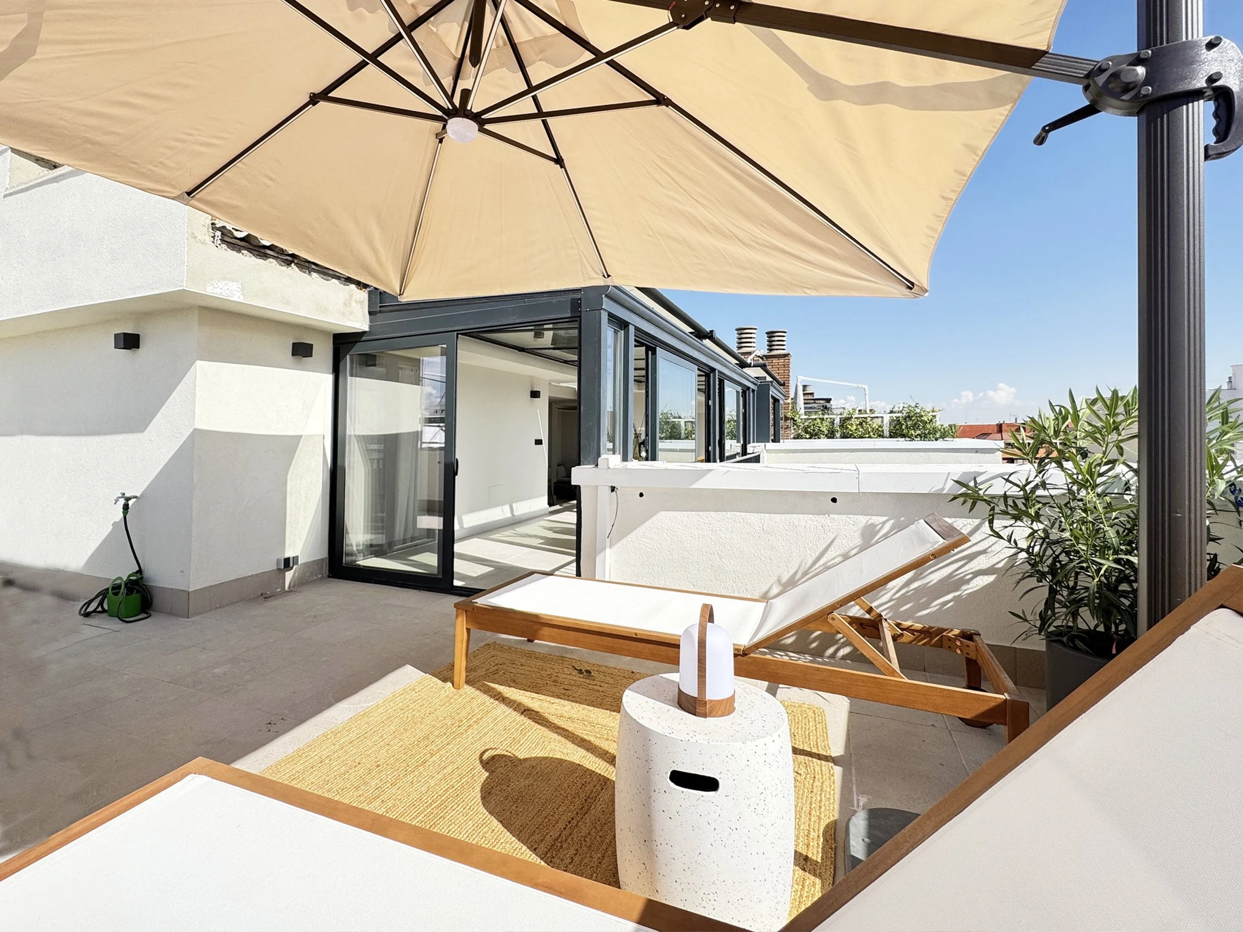 Terraza en un edificio con dos tumbonas de madera y tela blanca, una mesa pequeña con una lámpara y un paraguas grande beige, plantas en la esquina y puertas corredizas de cristal que conducen a un interior.