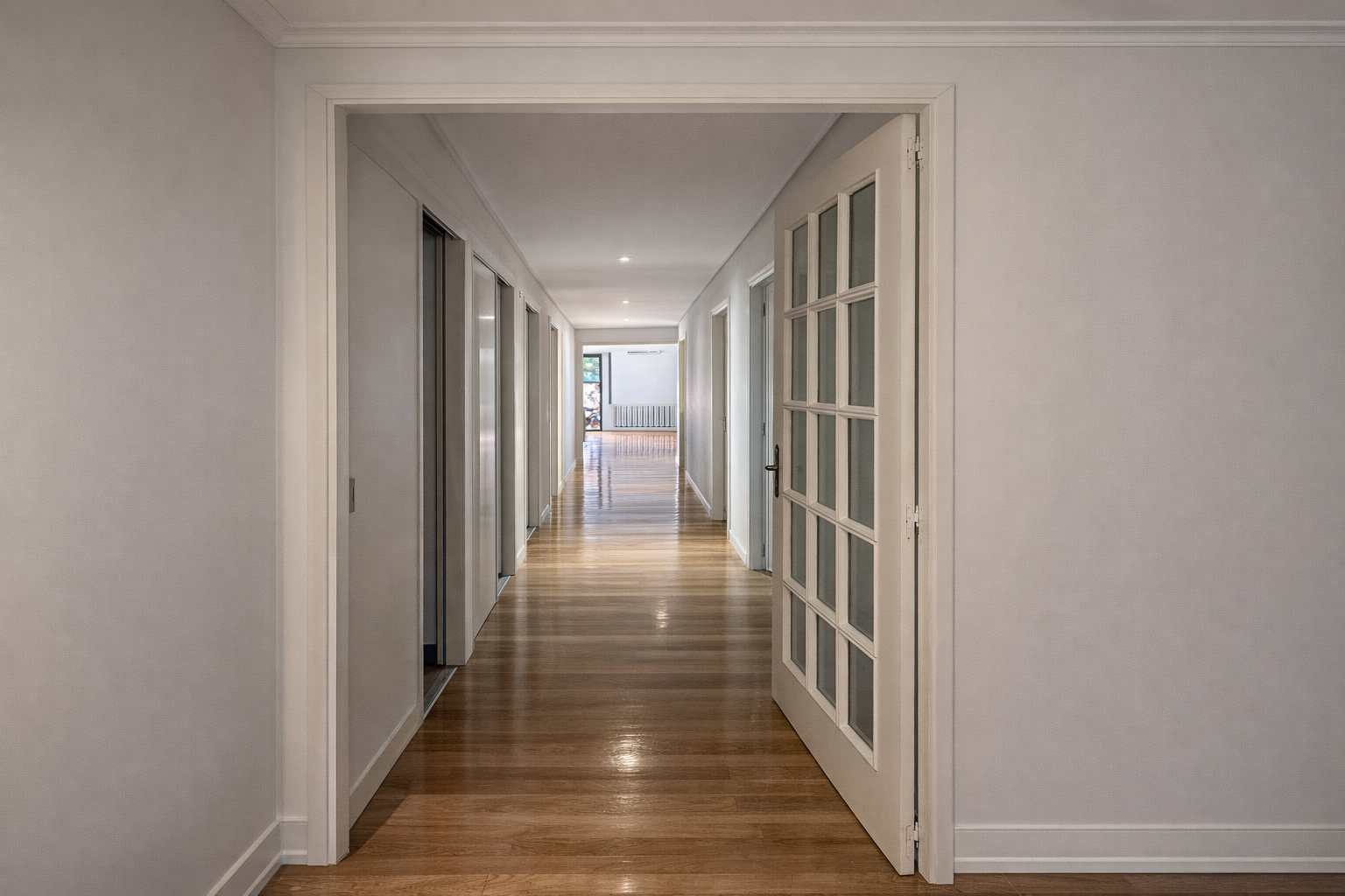 Pasillo interior de una casa con paredes blancas, suelo de madera y varias puertas de diferentes estilos.
