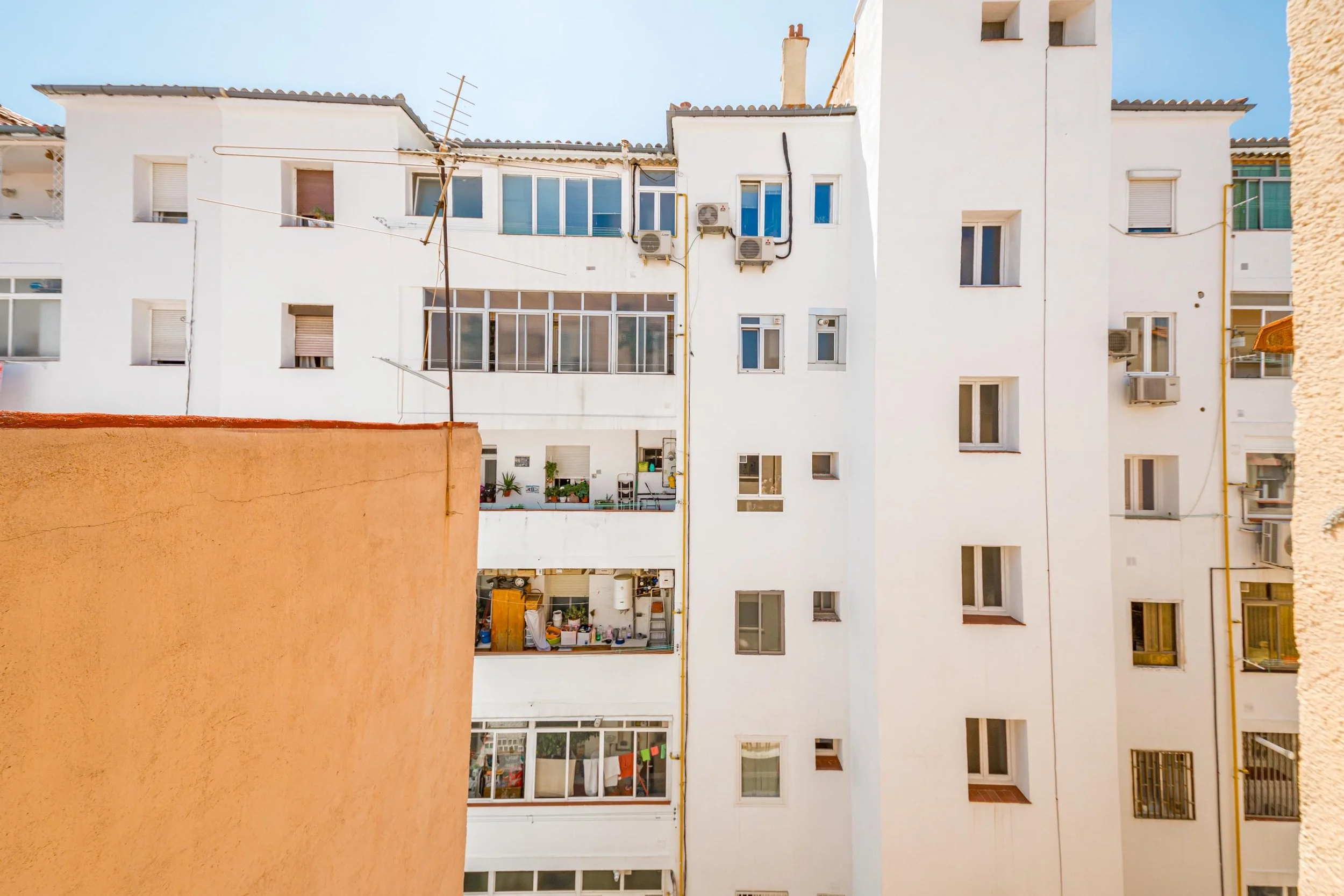 Vista de varios edificios residenciales blancos con ventanas y balcones, un muro naranja en primer plano y un cielo despejado.