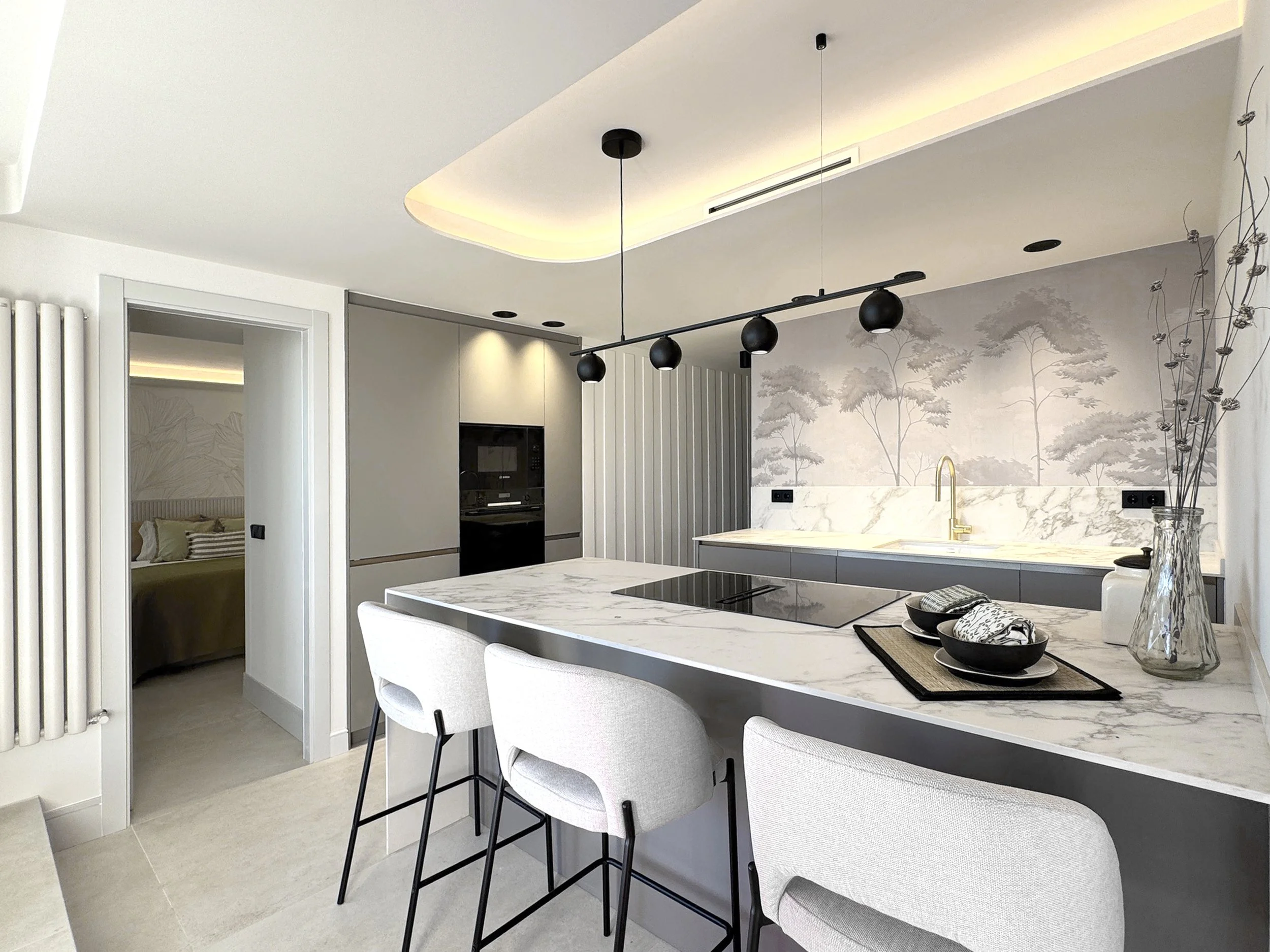 Vista de una cocina moderna con isla de piedra blanca, sillas blancas, paredes decoradas con papel tapiz de árboles y gabinetes grises, con un área de desayuno y espacios para electrodomésticos integrados.