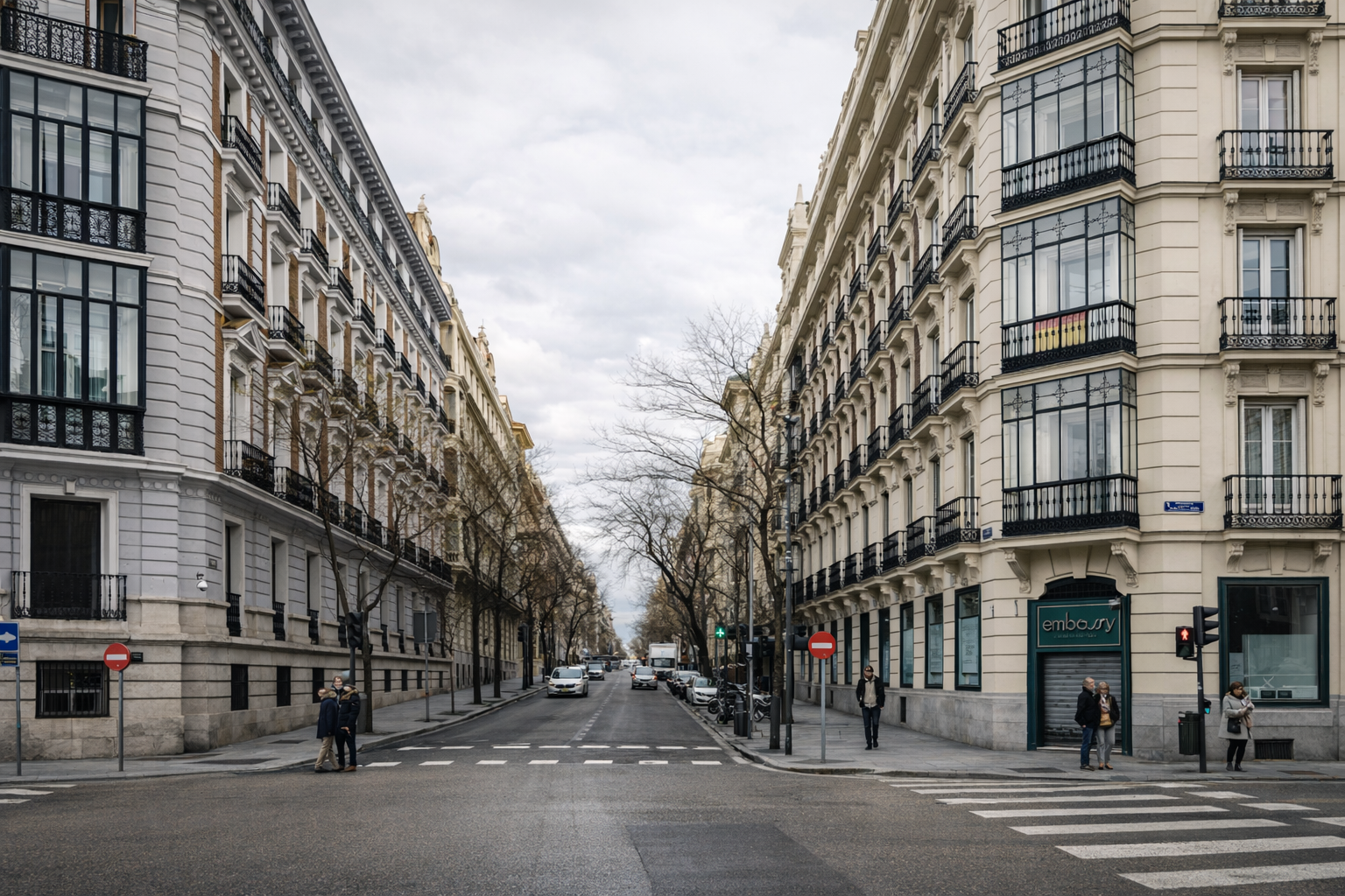 Vista de una calle en una ciudad con edificios de apartamentos de varios pisos en ambos lados, árboles sin hojas, semáforo en verde, varios peatones y autos en la calle, cielo nublado.