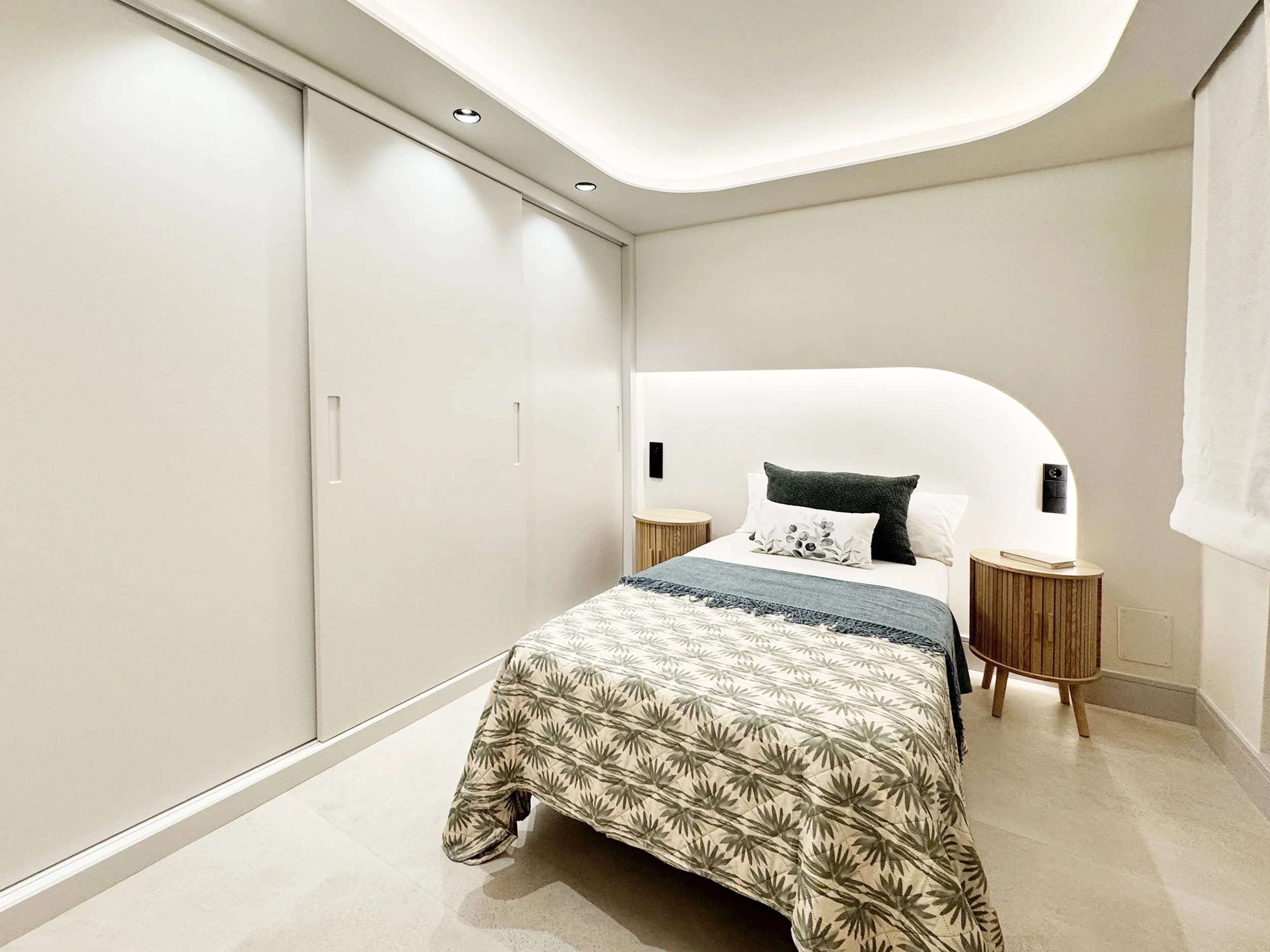 Habitación moderna con pared blanca, cama con ropa de cama con estampado de hojas, almohadas blancas y negras, mesas de noche de madera, iluminación suave y paneles con control de luz.