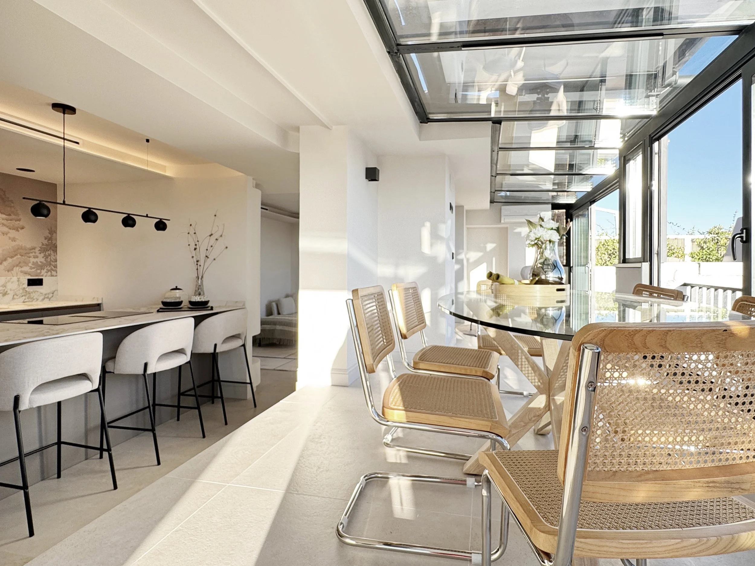 Cocina y comedor con mesas de madera y sillas, mucha luz natural, ventanas grandes con vista al exterior, decoraciones minimalistas con flores y vajilla.