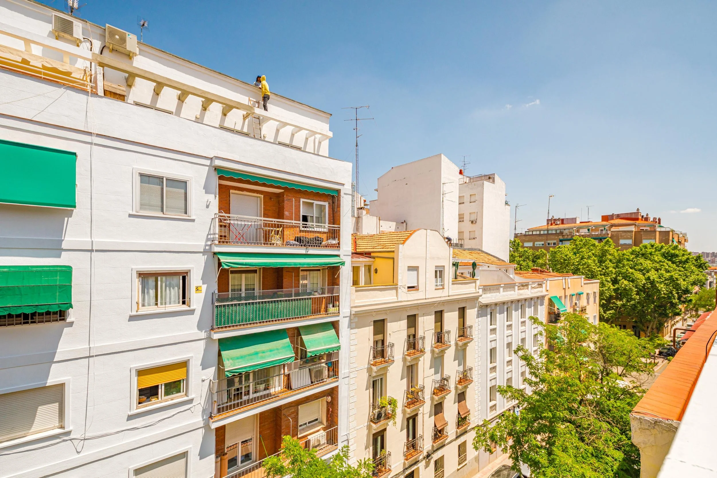 Edificios residenciales con balcones y ventanas, algunos con toldos verdes, y árboles frondosos en una ciudad bajo un cielo despejado.