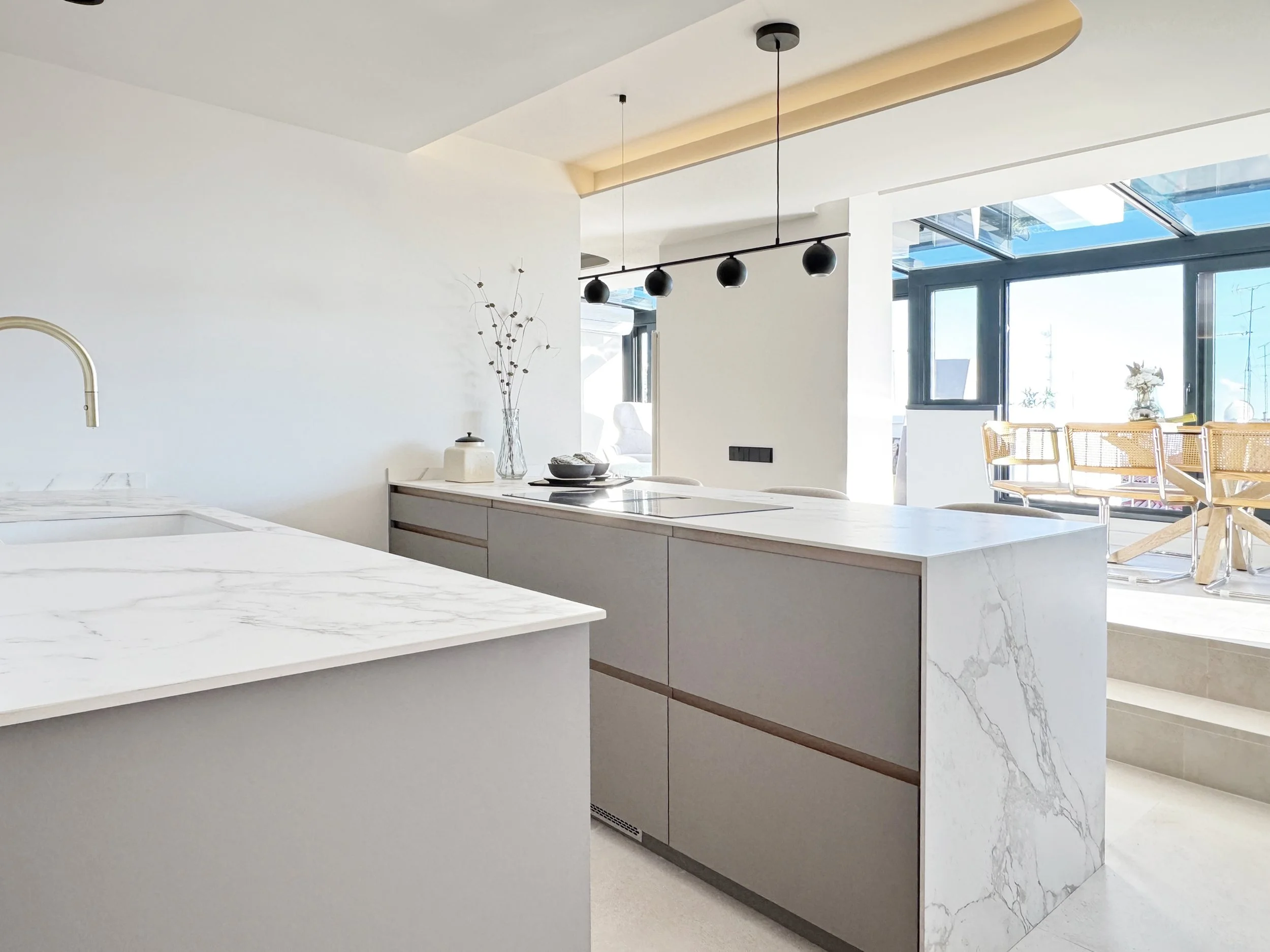 Cocina moderna con isla de mármol blanco y gabinetes beige; ventanas grandes con vista al balcón con sillas de madera.