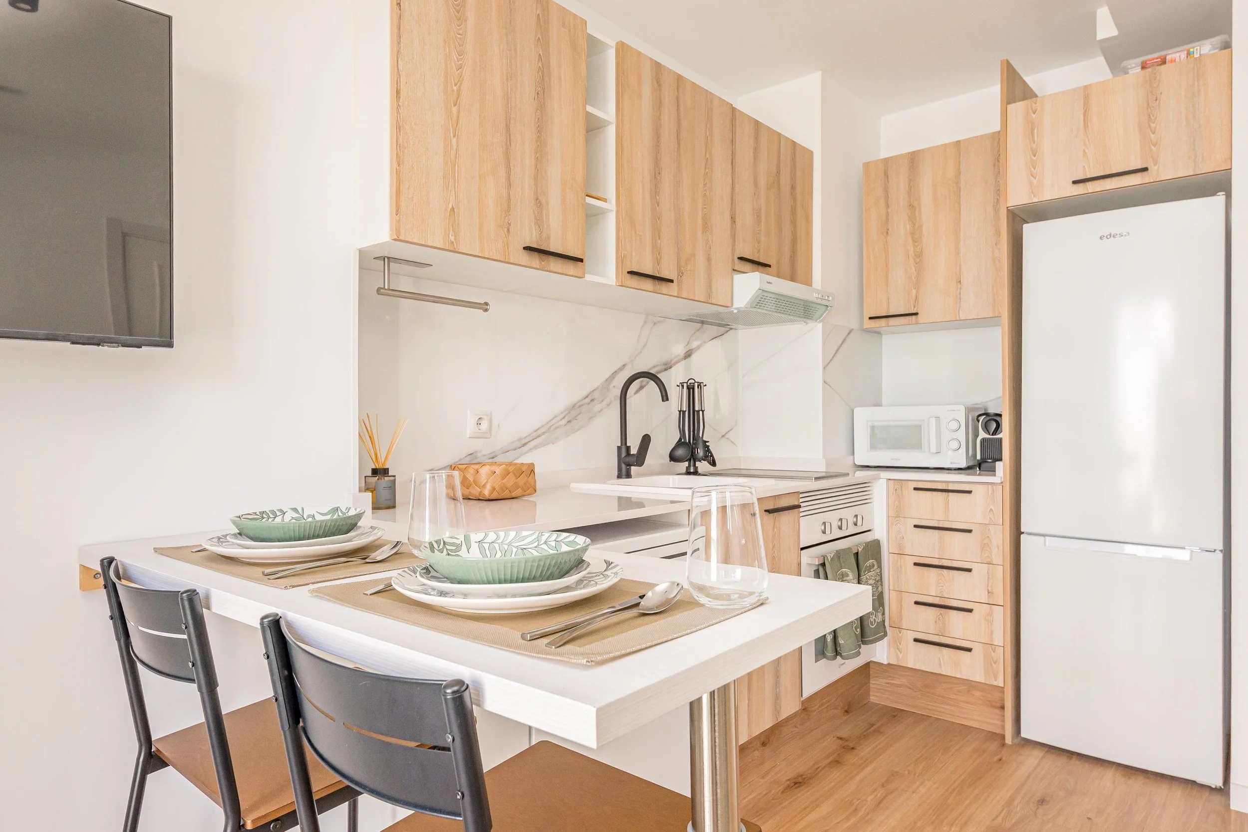 Cocina moderna con gabinetes de madera clara, electrodomésticos blancos, barra de desayunos con vajilla y cubiertos, suelo de madera.