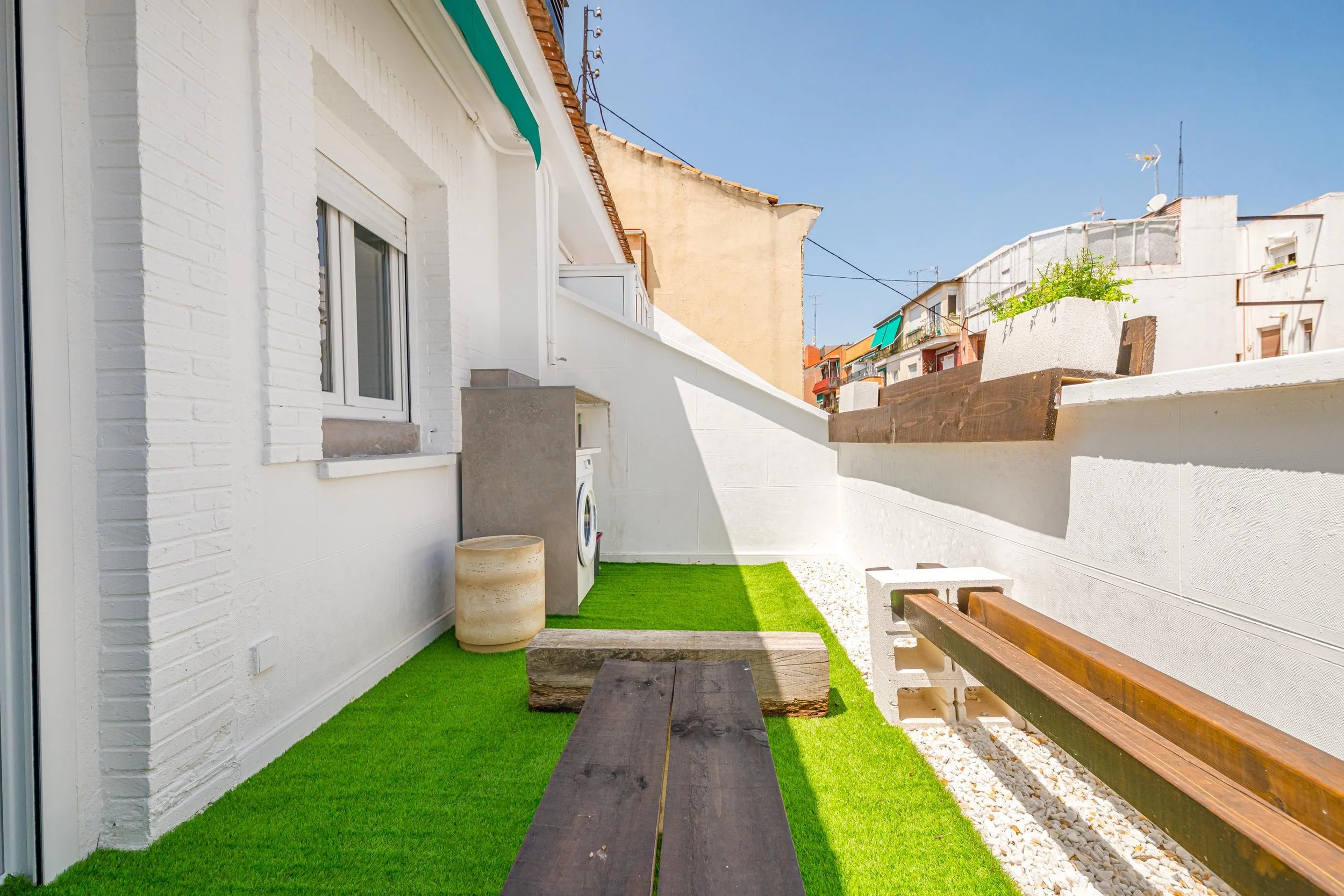 Balcón urbano con césped artificial, ventanas blancas, estructura de madera y una lavadora en un entorno residencial.