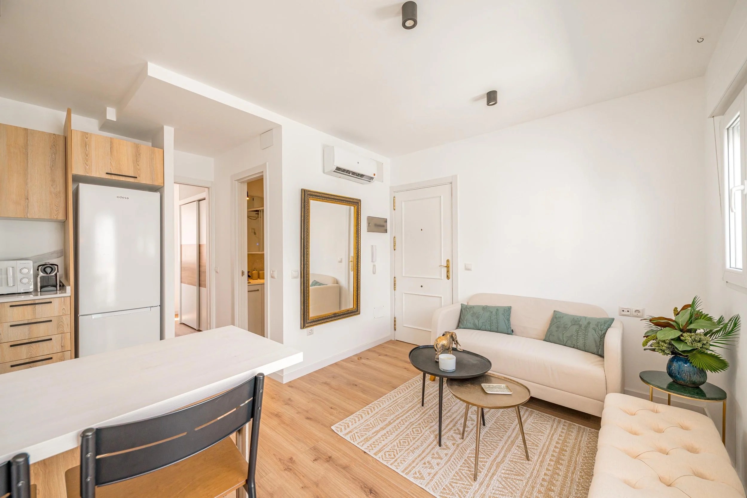 Sala de estar con sofá beige, mesas de centro negras y de madera, alfombra, plantas y pared blanca