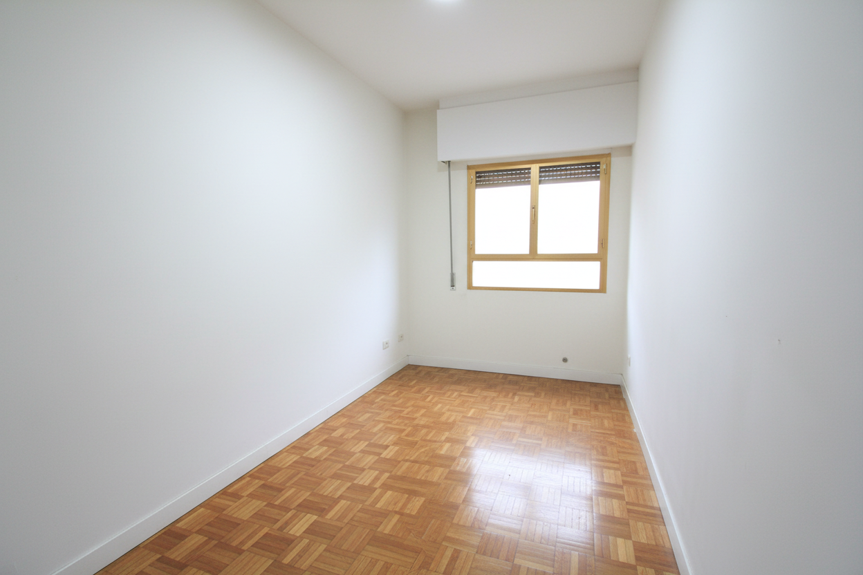 Una habitación vacía con paredes blancas, suelo de parquet, y una ventana con marco de madera y persiana enrollable.