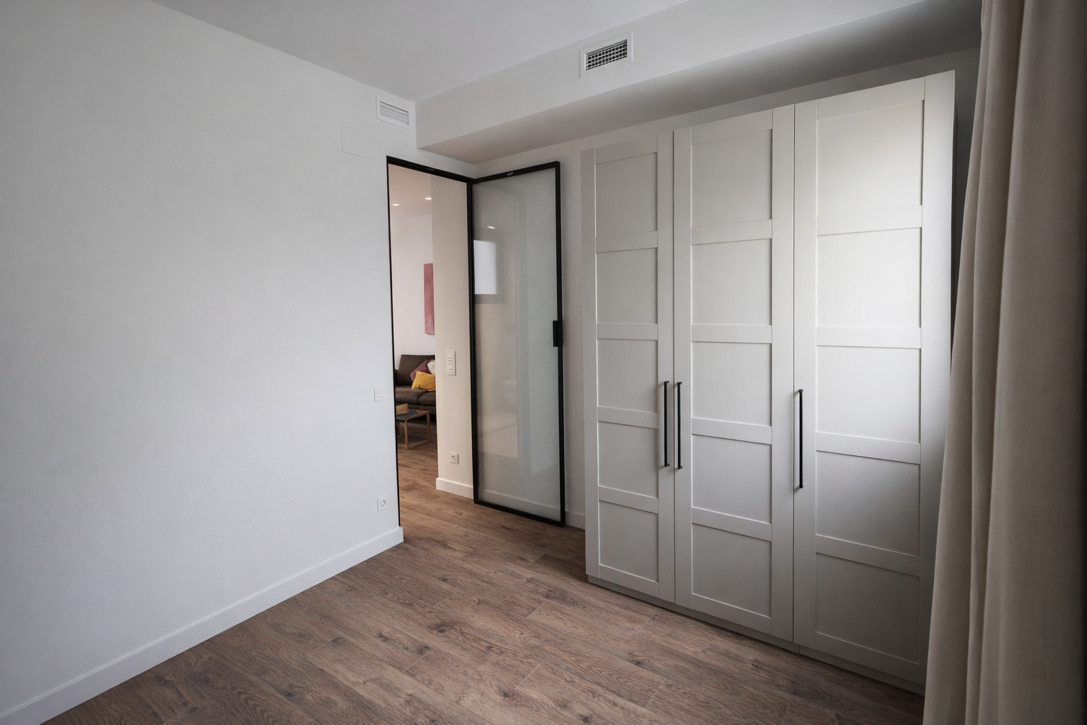 Habitación con armario de madera blanca y puerta de vidrio con marco negro, suelo de madera clara y cortina beige, vista hacia un área de sala con sofá y cojines en el fondo.