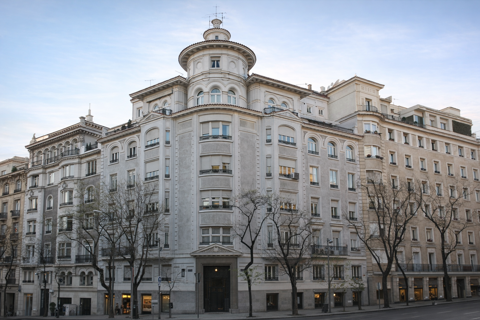 Edificio de estilo clásico con varias plantas, balcones y ventanas grandes, árboles en la acera y cielo despejado.
