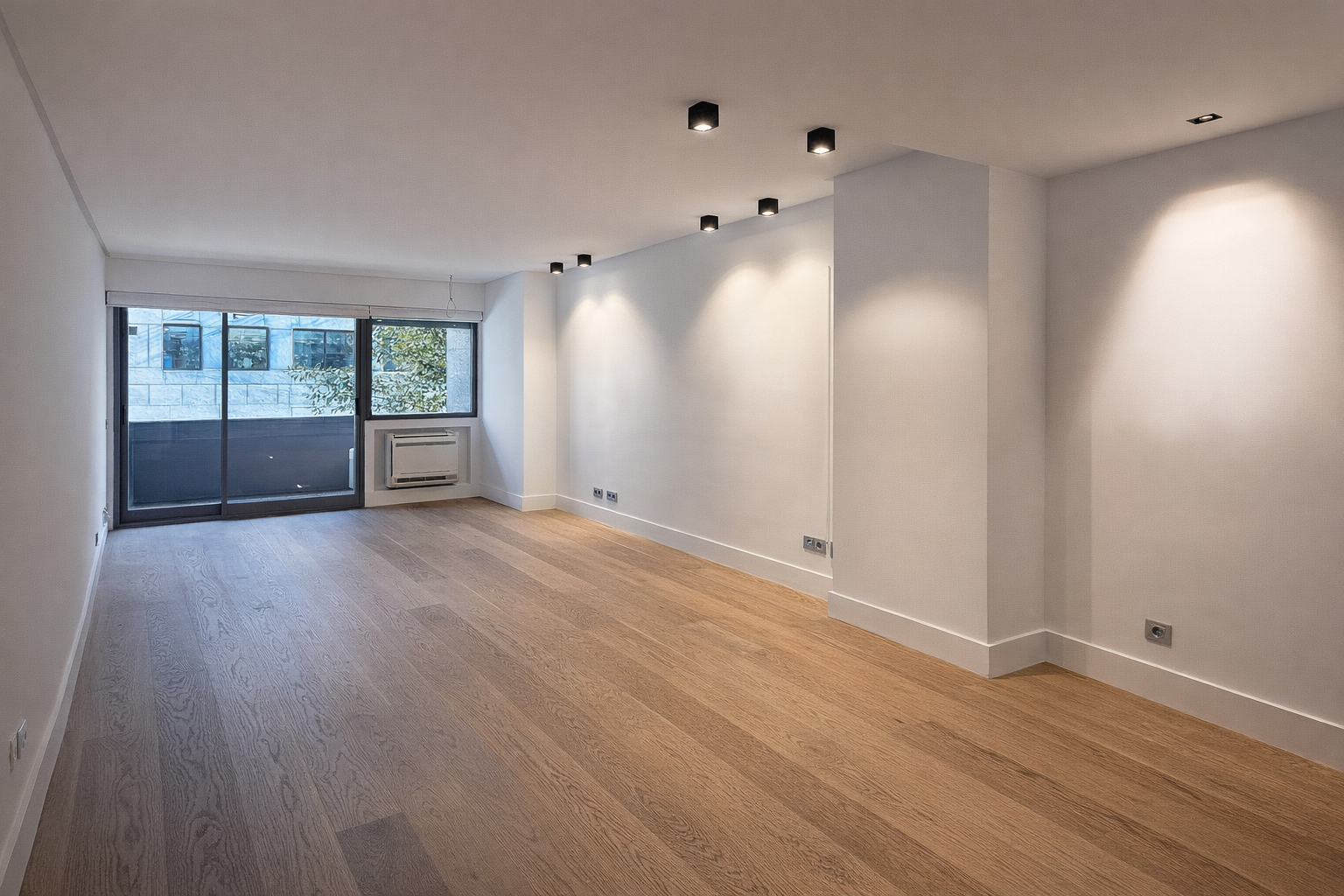Sala vacía con paredes blancas, suelo de madera, ventanas amplias, aire acondicionado, y luces empotradas en el techo.