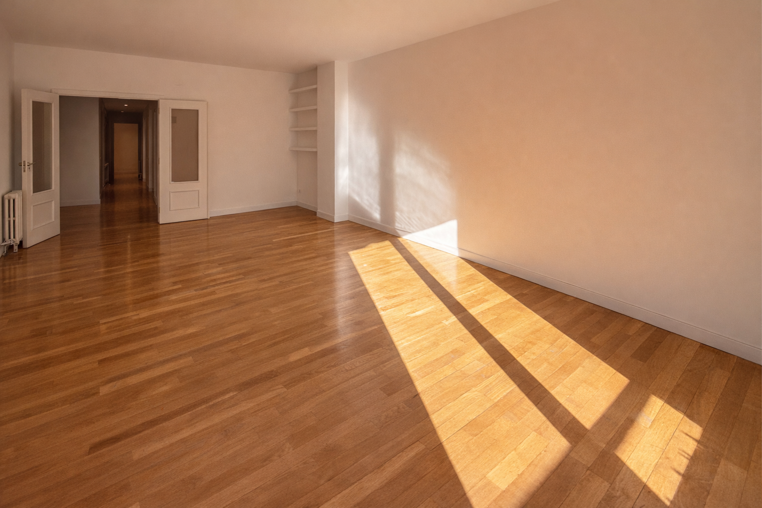 Sala vacía con paredes blancas, piso de madera y puertas abiertas a un pasillo.