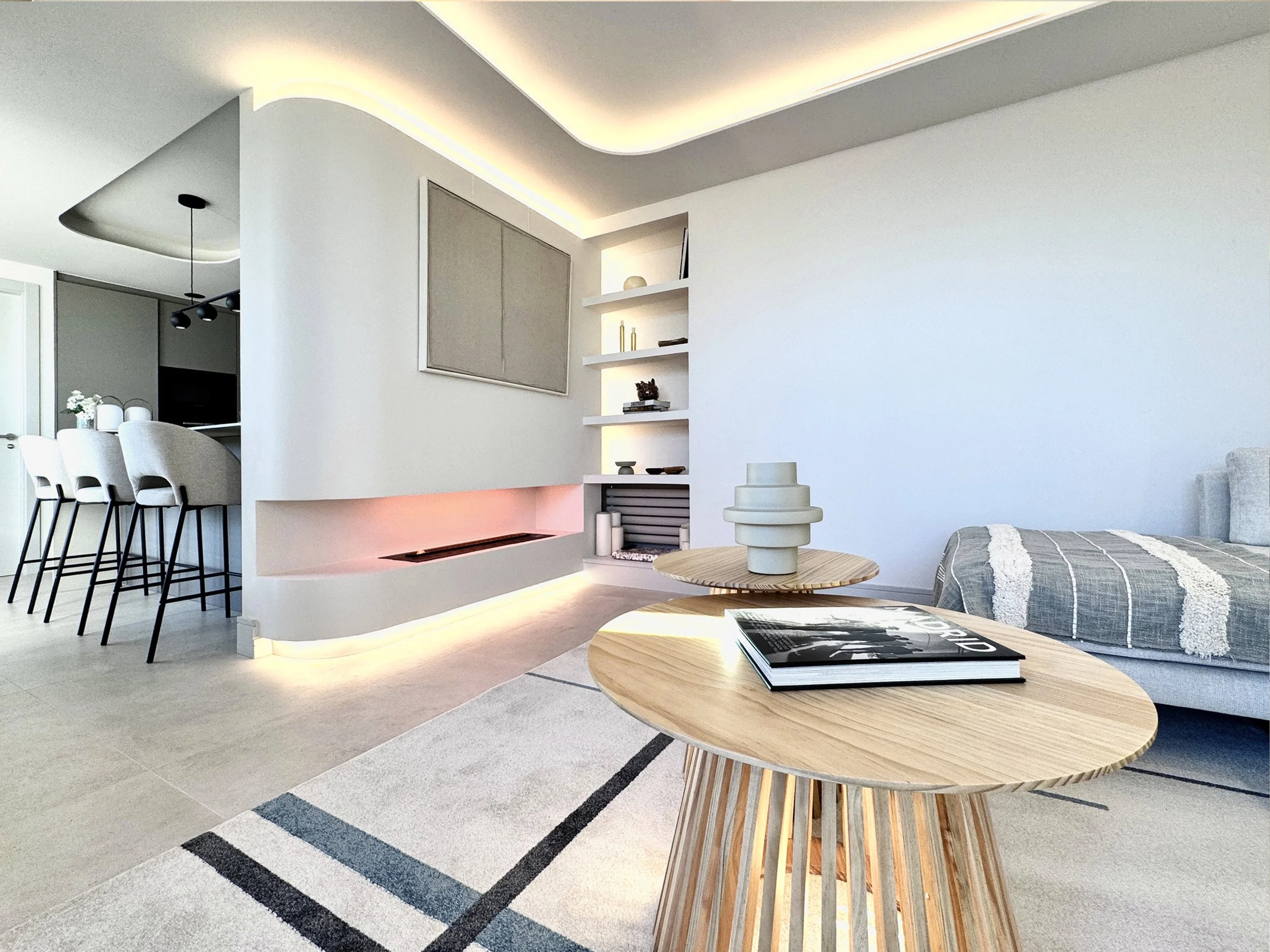 Sala moderna con mesas de madera, una pared blanca con estanterías y decoración minimalista, y un área de comedor en el fondo con sillas altas y un espacio de televisión.