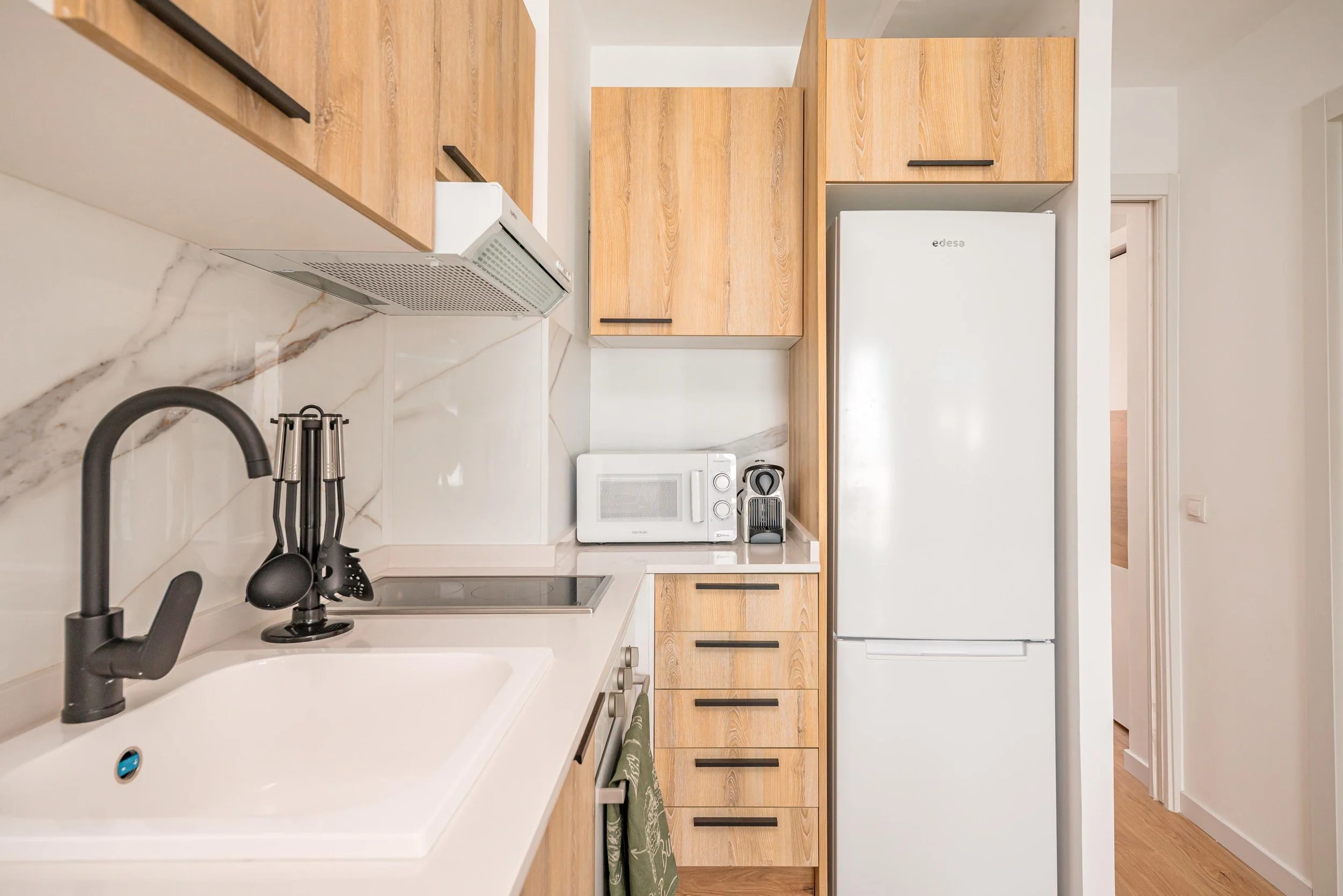 Cocina moderna con gabinetes de madera clara y electrodomésticos blancos, incluyendo refrigerador, microondas, estufa eléctrica y campana extractora, en un espacio compacto y ordenado.