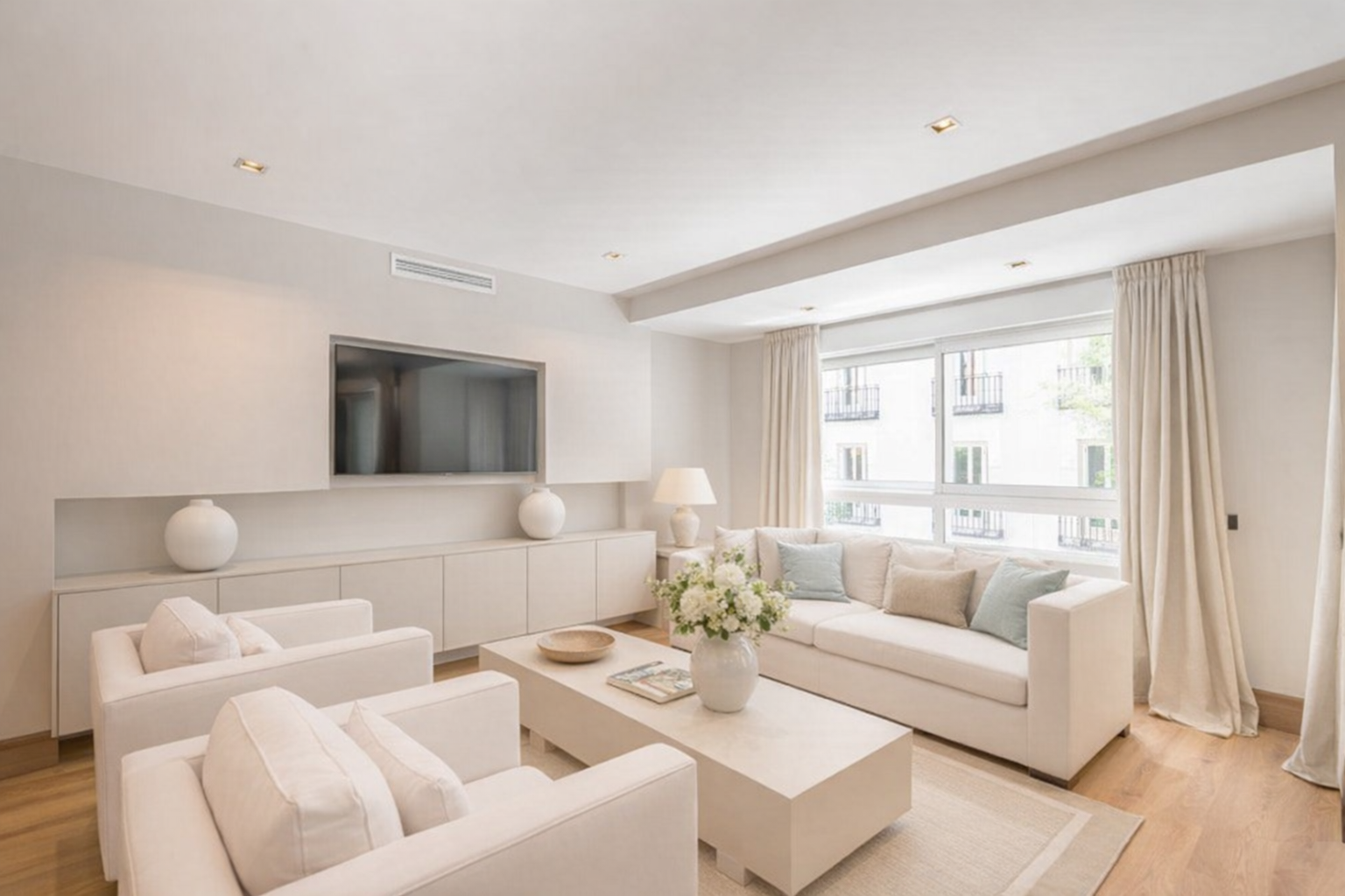 Sala de estar luminosa con muebles blancos, sillones, una mesa de centro, un sofá, cortinas claras y ventanas grandes con vista a un edificio