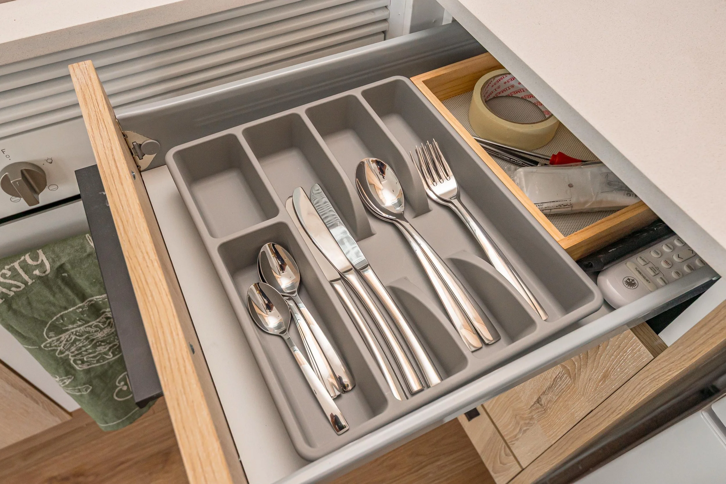 Cajón de cocina con cubiertos de acero inoxidable, incluyendo cucharas, tenedores y cuchillos, organizado en compartimentos de una nuez. Hay cinta adhesiva y herramientas en la bandeja superior, y una televisión remota en la parte inferior derecha.