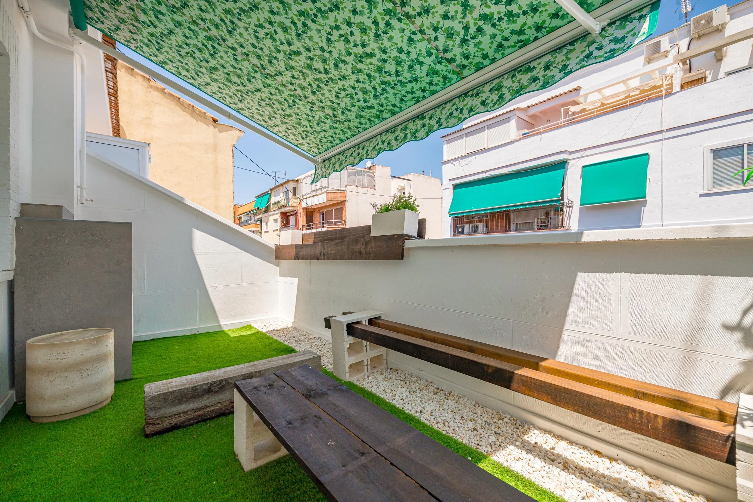 Pequeño patio con césped artificial, bancos de madera, y paredes blancas con maceta en la parte superior, con edificios de fondo y sombrilla verde de jardín.