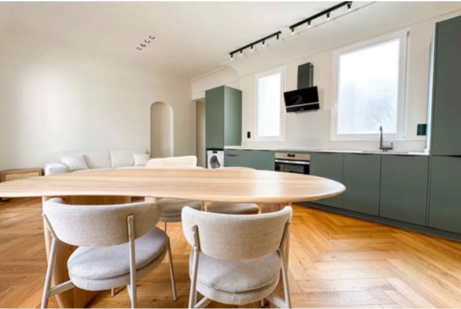 Cocina moderna con gabinetes verdes, horno, campana extractora, fregadero, lavadora, ventanas grandes, comedor con mesa de madera ovalada y sillas acolchadas, suelo de parquet de madera.