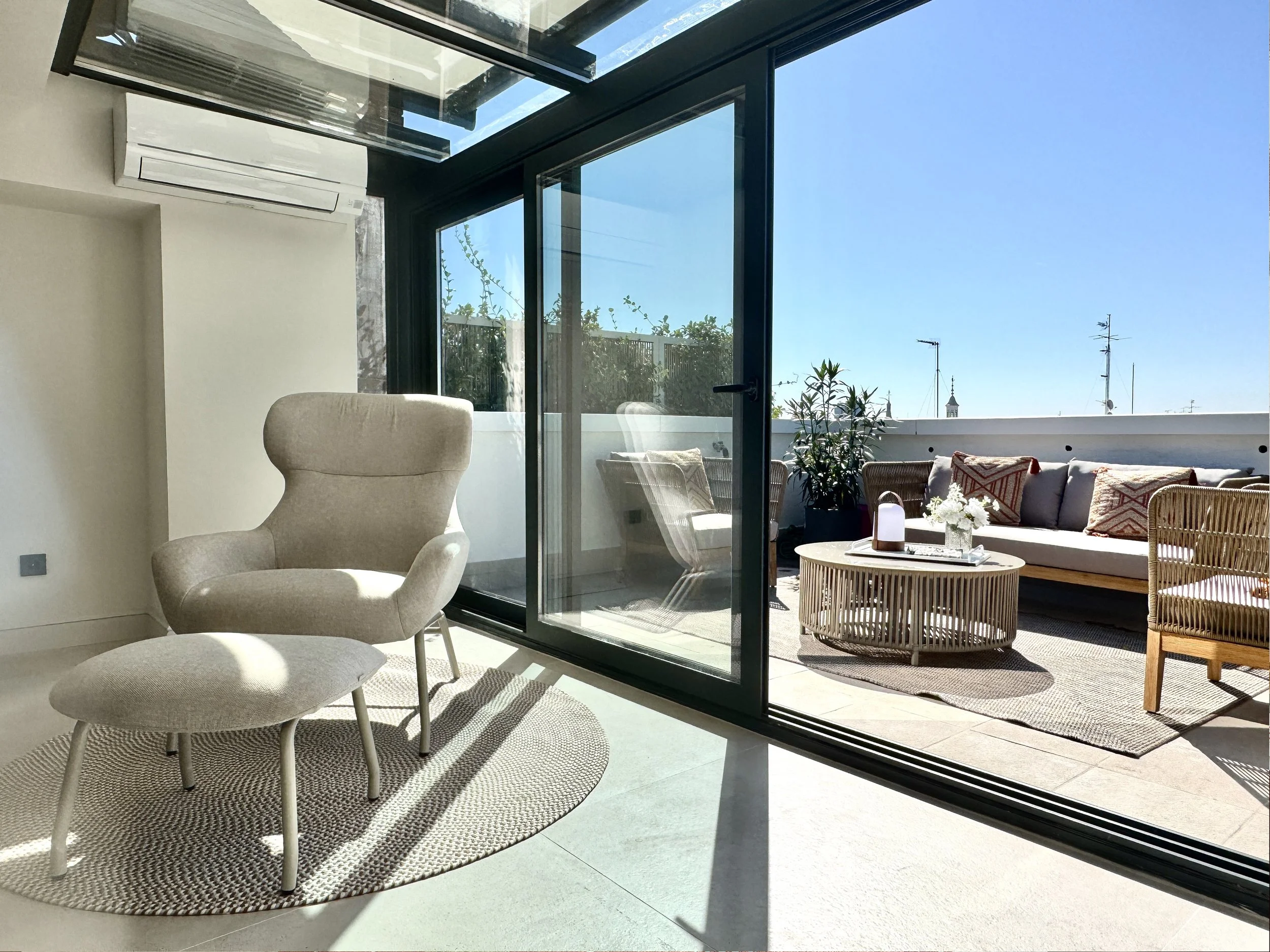 Sala moderna con silla y ottoman cerca de puertas de cristal que abren a una terraza con sillones, mesa de café y plantas bajo cielo despejado.