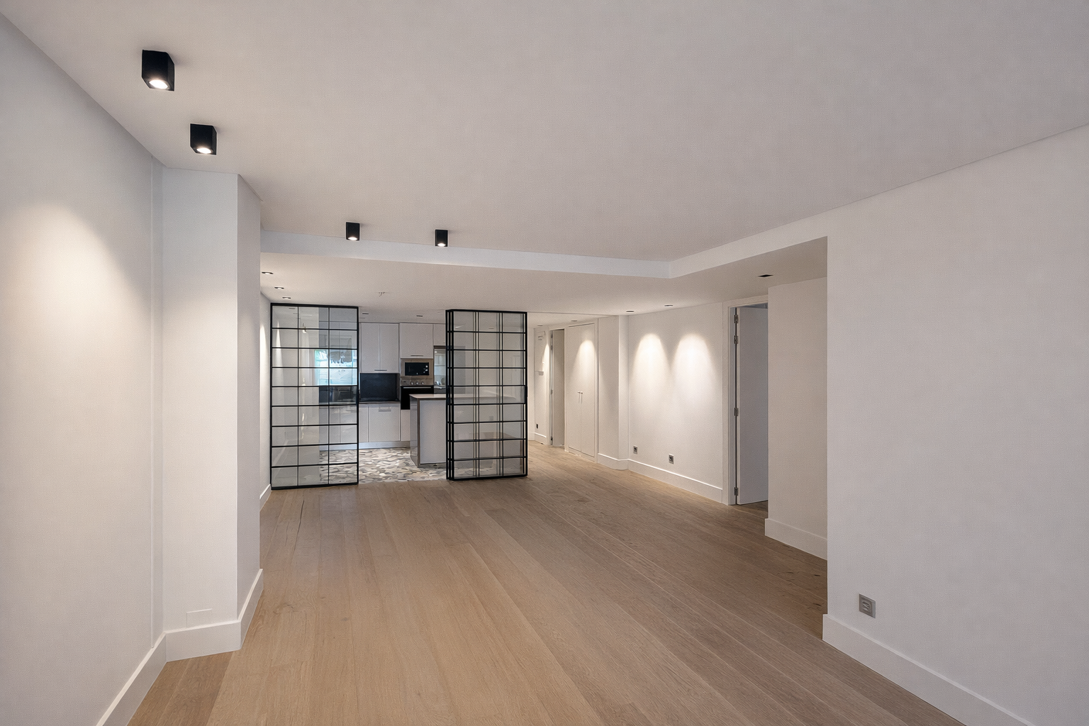 Sala vacía con paredes blancas, suelo de madera clara, división de vidrio negro y cocina moderna al fondo.