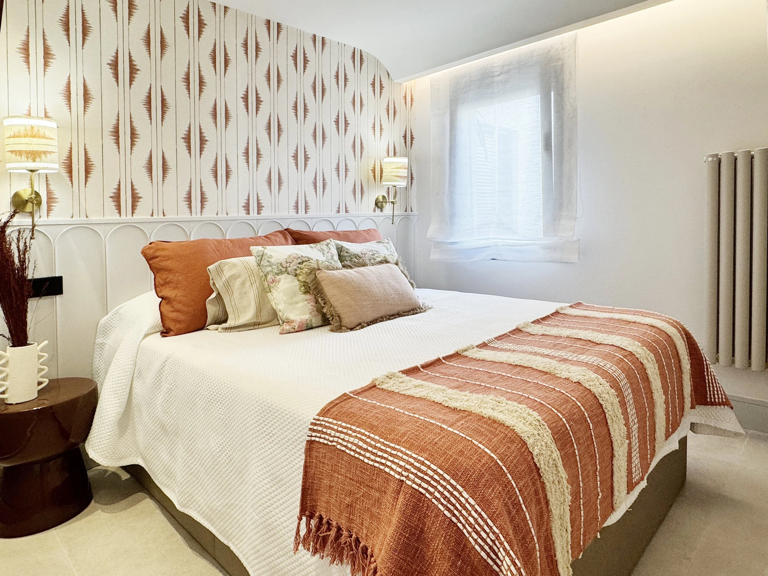 Habitación con cama decorada con almohadas en tonos terracota y un cobertor a rayas en colores similares, pared con papel tapiz de diseño geométrico en tonos marrón, lámparas de pared, una ventana con cortina blanca, y un radiador en la esquina derec