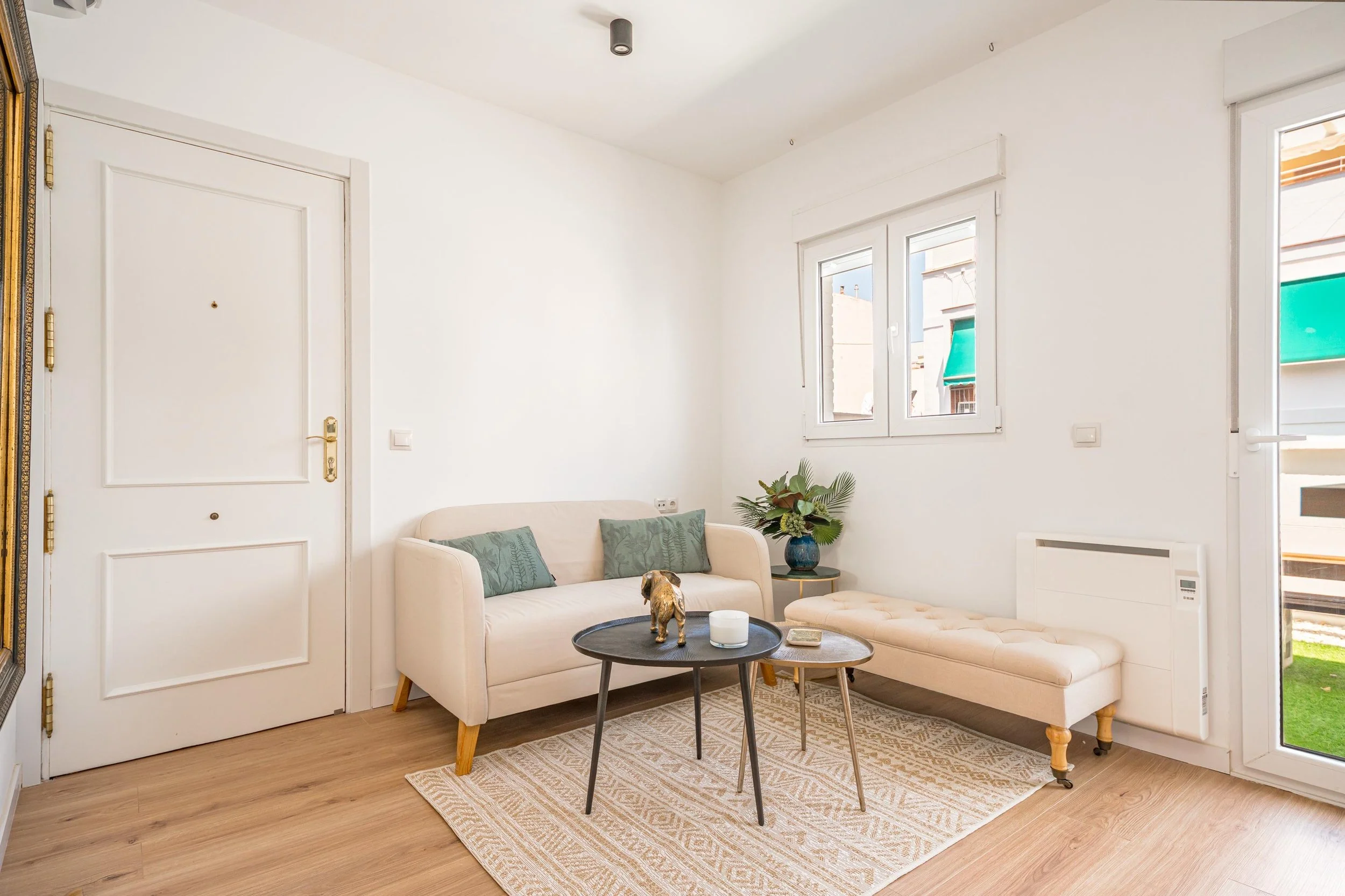 Sala de estar con sofá beige, mesa de centro redonda negra, una banca beige y una planta en una esquina, ventanas con vista a edificios cercanos, piso de madera y paredes blancas.