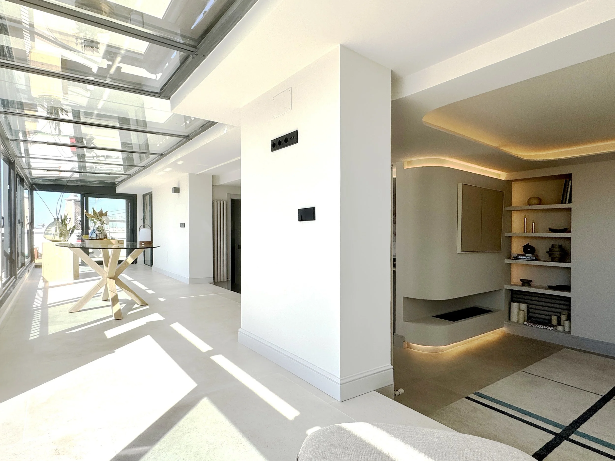 Interior de una casa moderna con paredes blancas, techo de vidrio y muebles de madera clara. Hay una mesa decorativa con jarrones y plantas en el espacio principal, y una zona con repisas y una chimenea en una esquina.