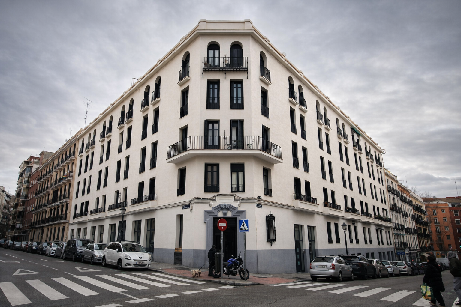 Edificio de color blanco con varias ventanas y balcones en la esquina de una calle en un día nublado.