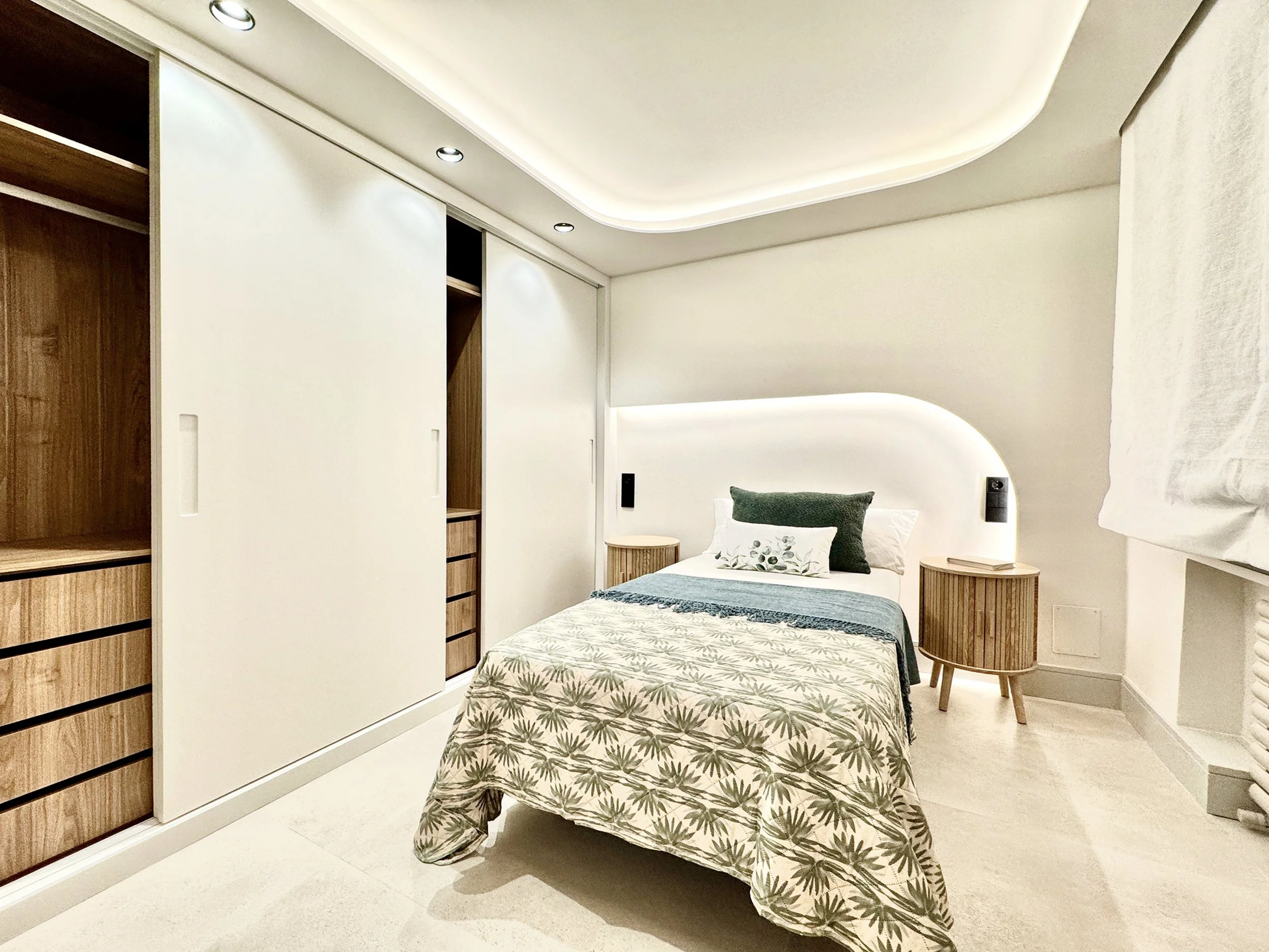 Habitación moderna con cama doble, cabecera curva con iluminación LED, accesorios de madera, paredes blancas, y cortinas blancas.