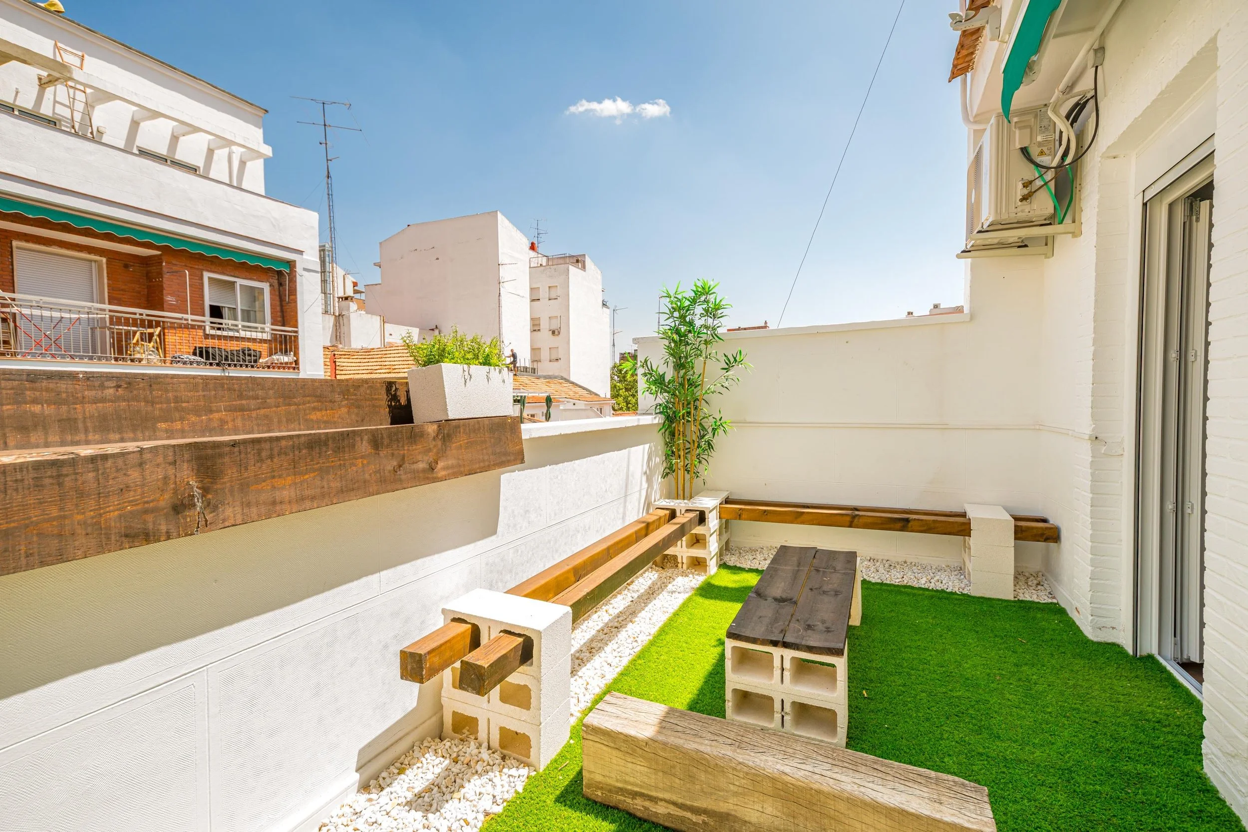 Terraza de un apartamento con piso de césped artificial, paredes blancas y bancos de madera suspendidos en bloques de cemento, con plantas y edificios de fondo bajo un cielo despejado.