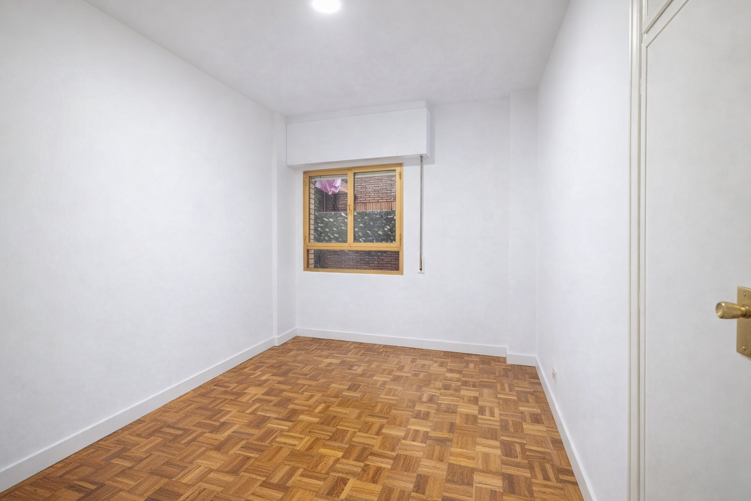 Habitación vacía con paredes blancas, suelo de madera y ventana con vista a un edificio de ladrillos.