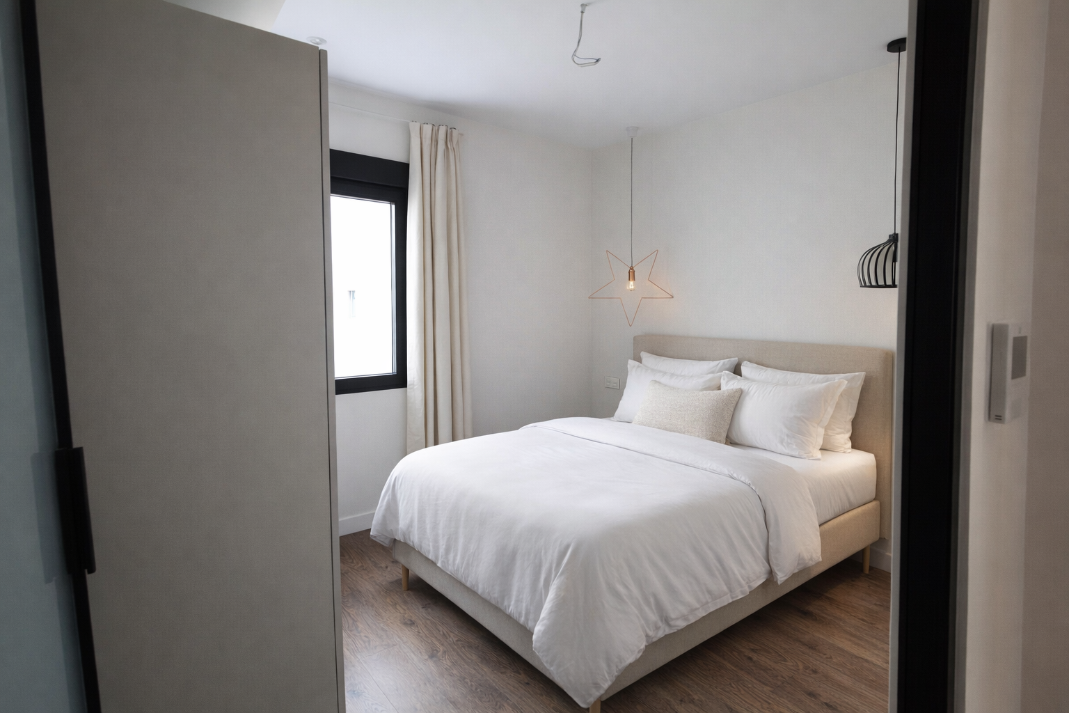 Habitación moderna con cama grande, blanca y ropa de cama, ventana con cortinas claras, lámpara de diseño en forma de estrella y espacio ordenado y luminoso.