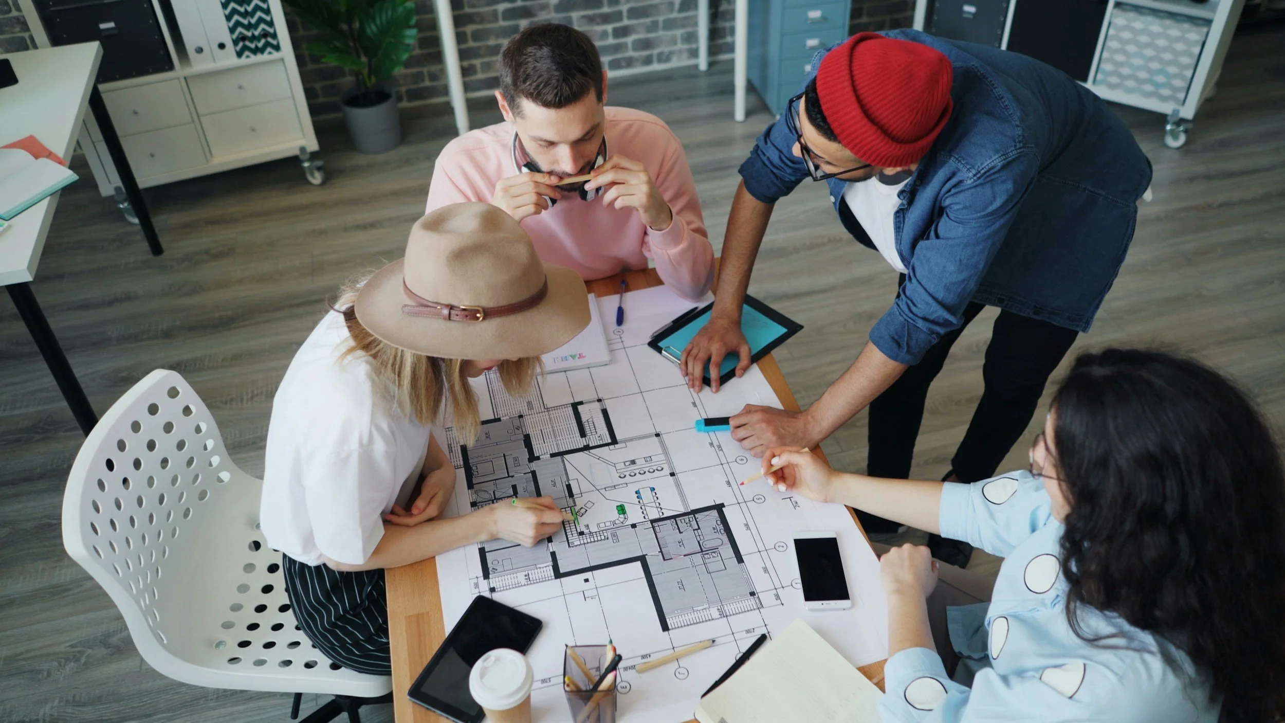 Group of five people collaborating around a table with architectural floor plans, discussing and analyzing the designs in an office environment.