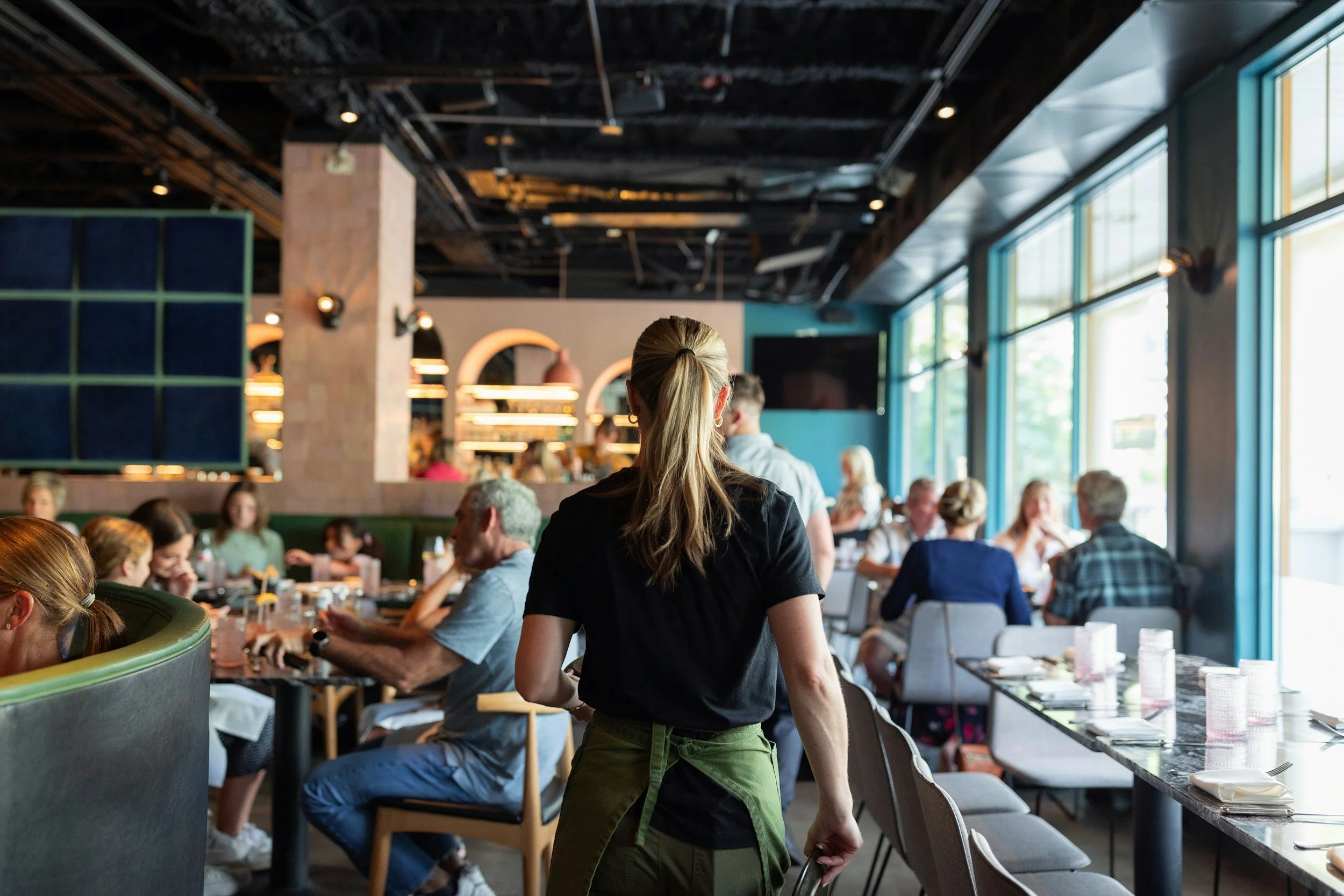 A busy restaurant with people seated at tables, a woman with blonde hair tied back in a ponytail standing near a table, and large windows letting in natural light.