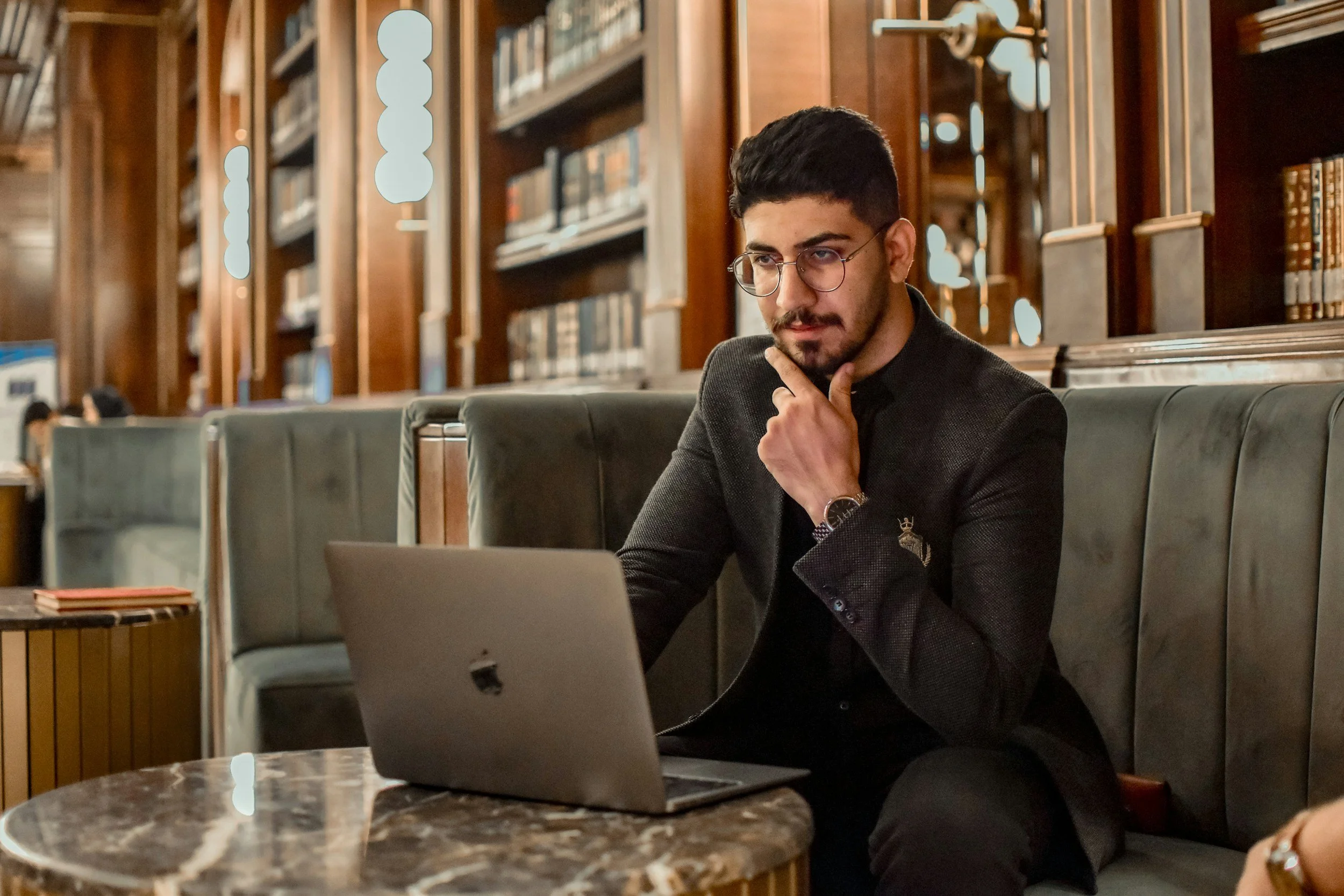 A man with dark hair and glasses, wearing a dark blazer, sitting at a table with a laptop, in a library with wooden shelves filled with books.
