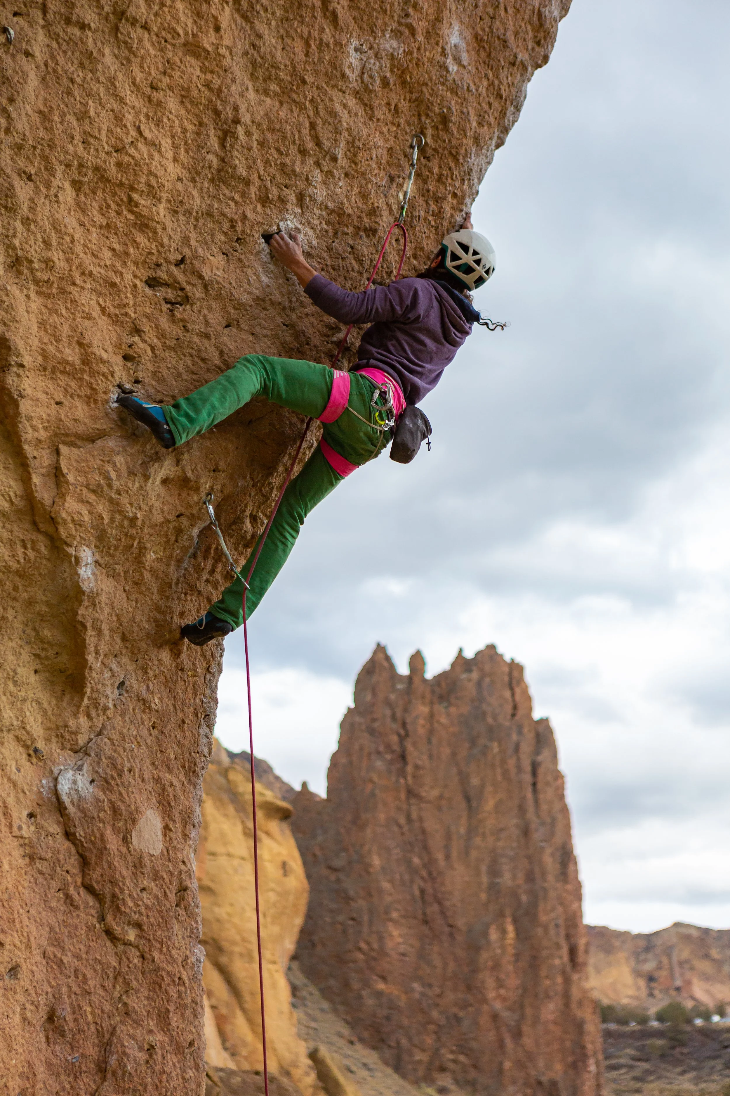A person wearing green pants, a bright pink rock climbing harness, a purple sweatshirt and a white helmet is rock climbing at Smith Rock Park. There is another rock peak in the distance.