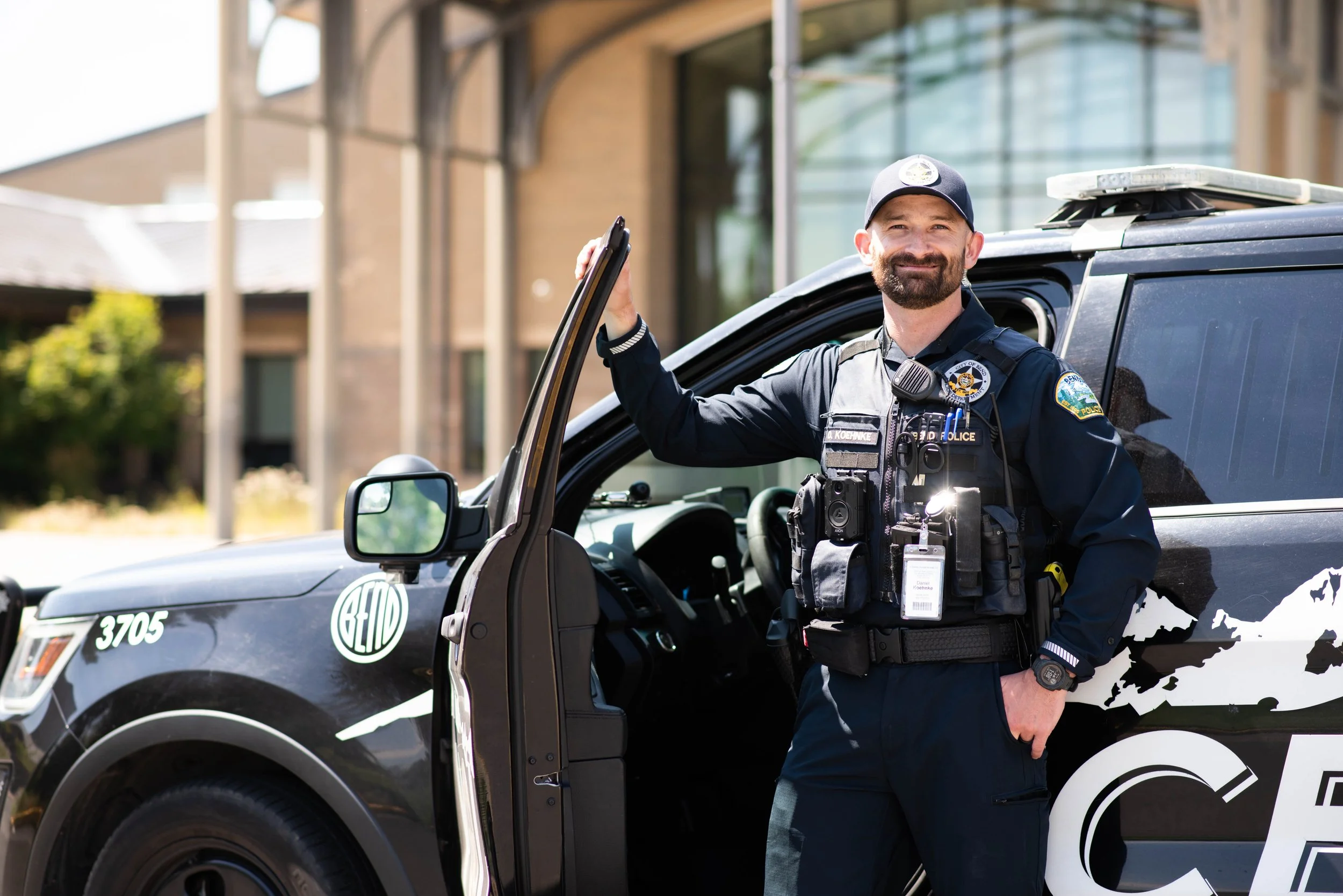 A police officer standing next to a police vehicle with the door open, smiling and looking at the camera.