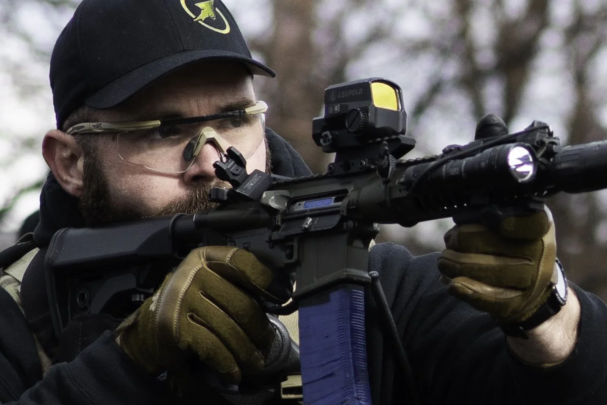 A man wearing tactical gear, protective glasses, and a black cap aiming a rifle with a scope outdoors.