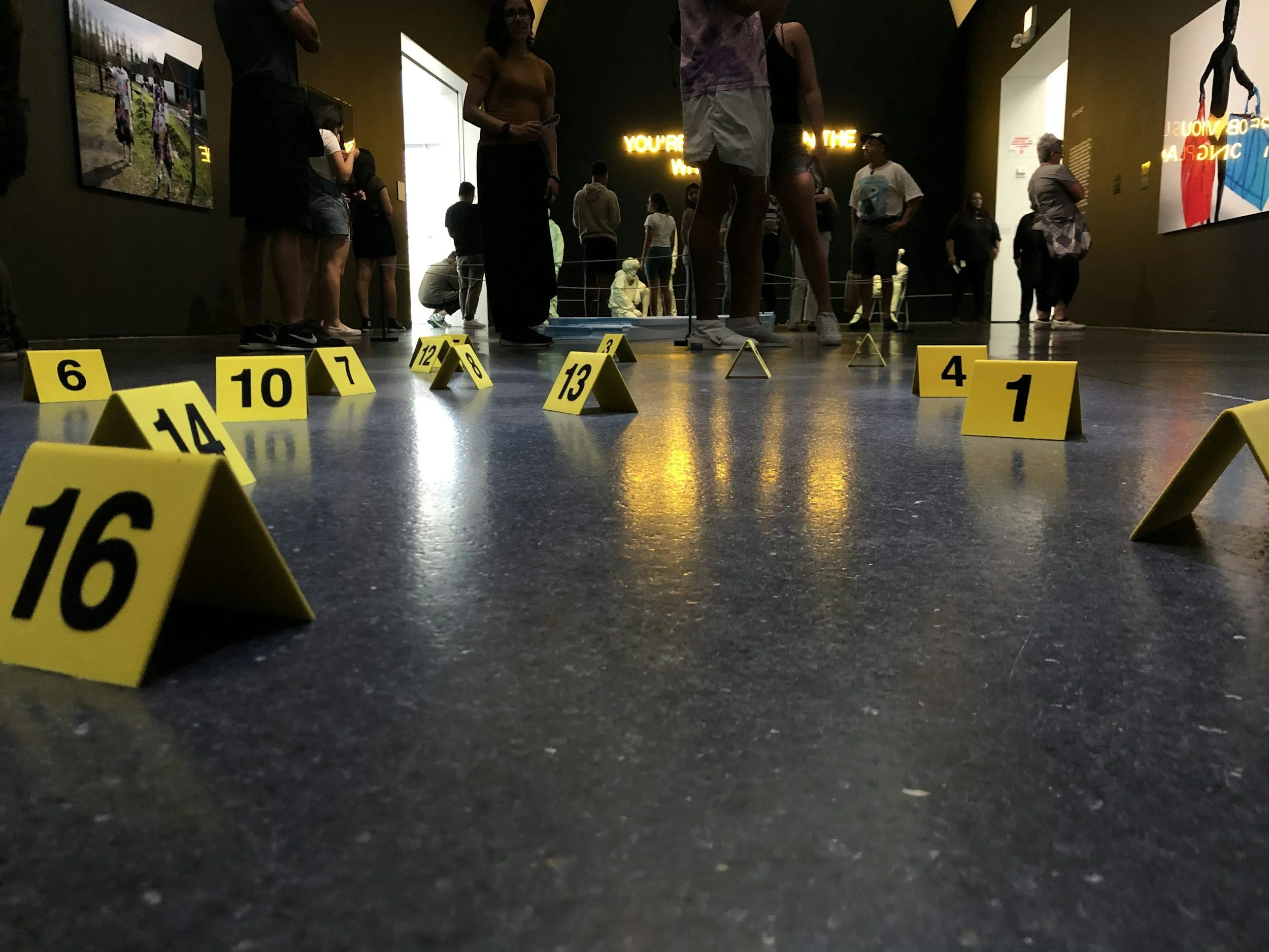 Indoor exhibit with yellow evidence markers numbered 1 to 16 scattered on the floor, and people observing and walking around in the background.