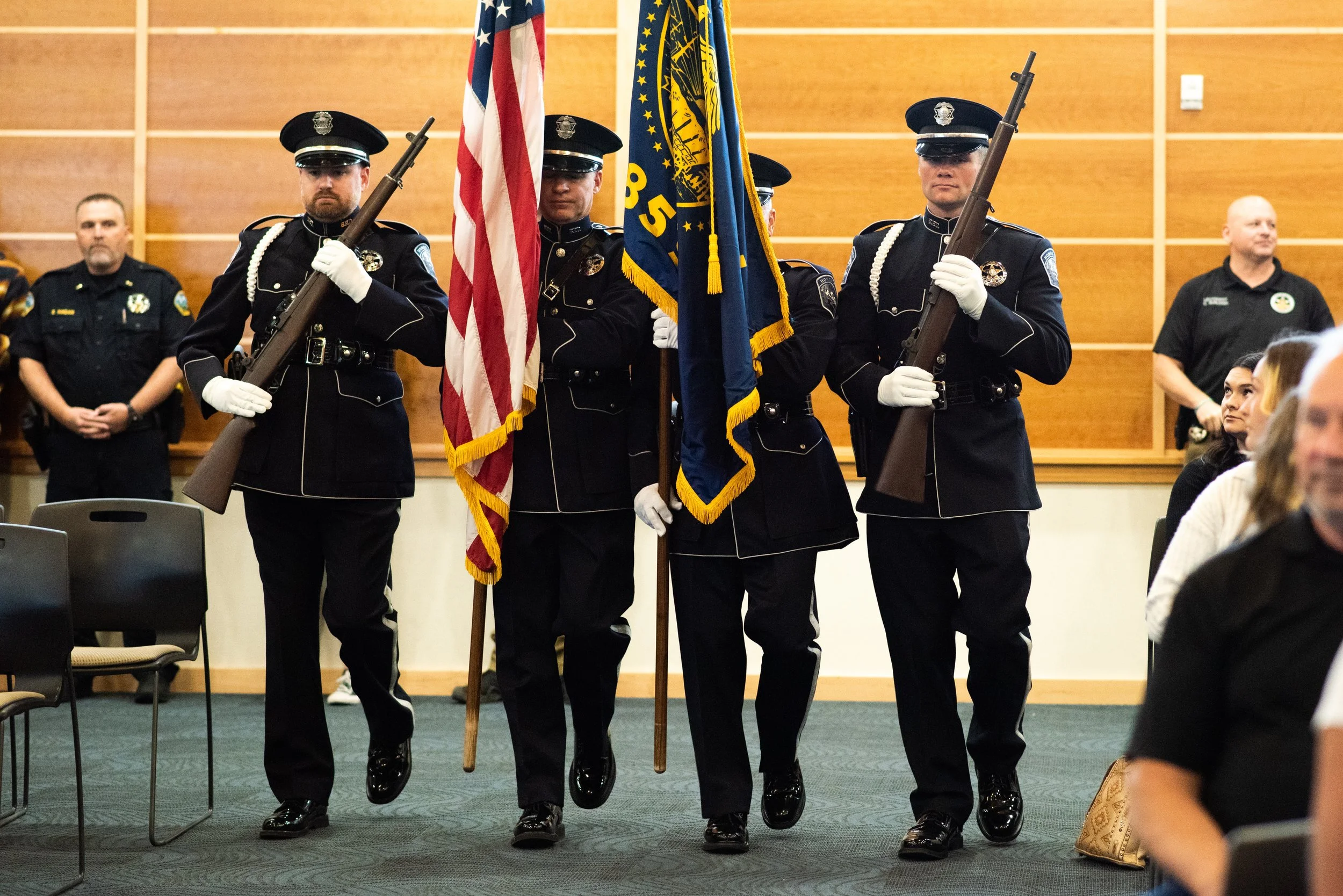 Police officers in ceremonial uniforms carrying flags during an indoor procession, with some seated audience watching.